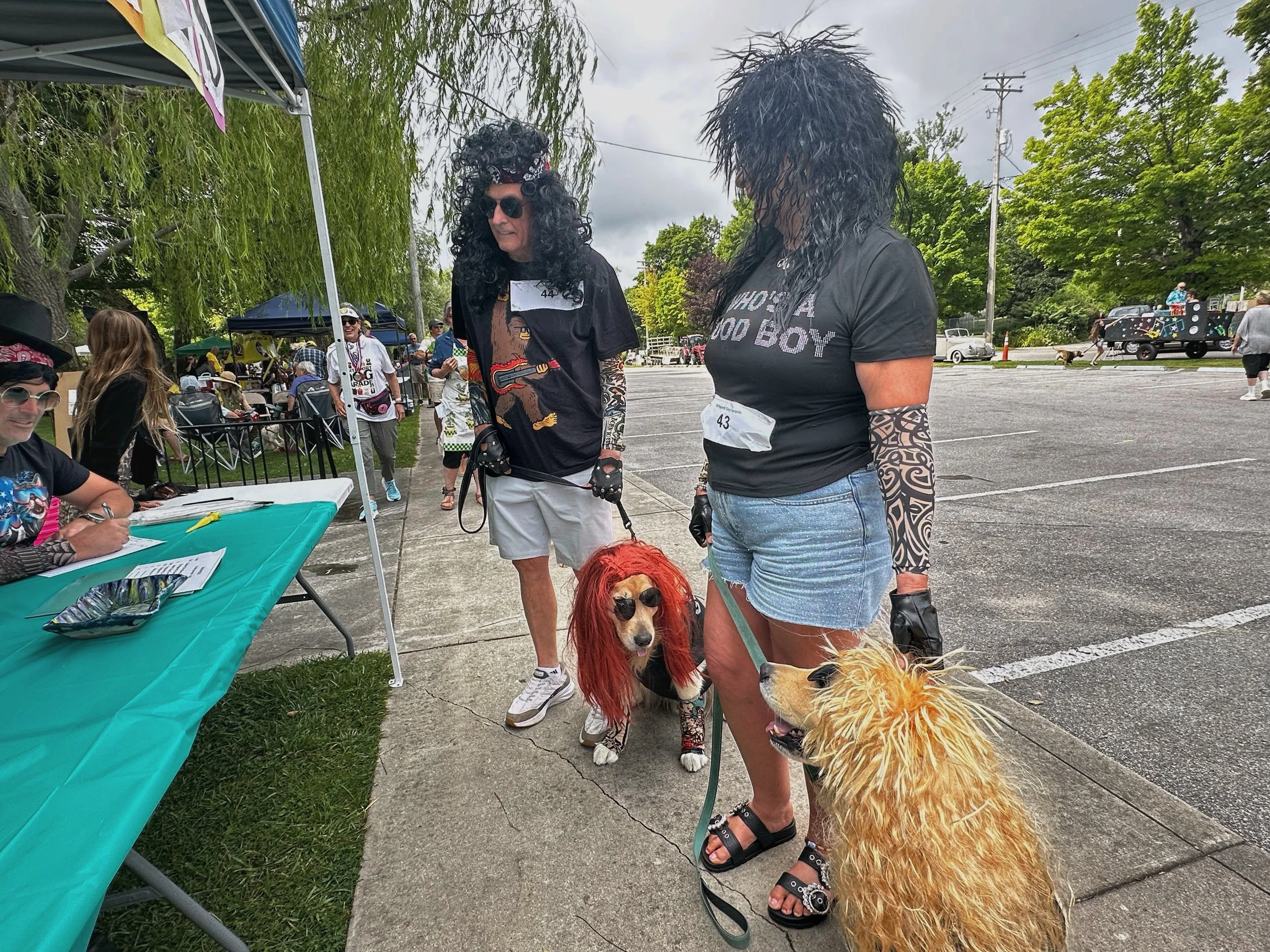 Group of people, some dressed in 1980s rock star costumes, gathered outdoors for an event. Two women in the center are wearing black wigs and sunglasses, and a dog with a lion costume is in the foreground.