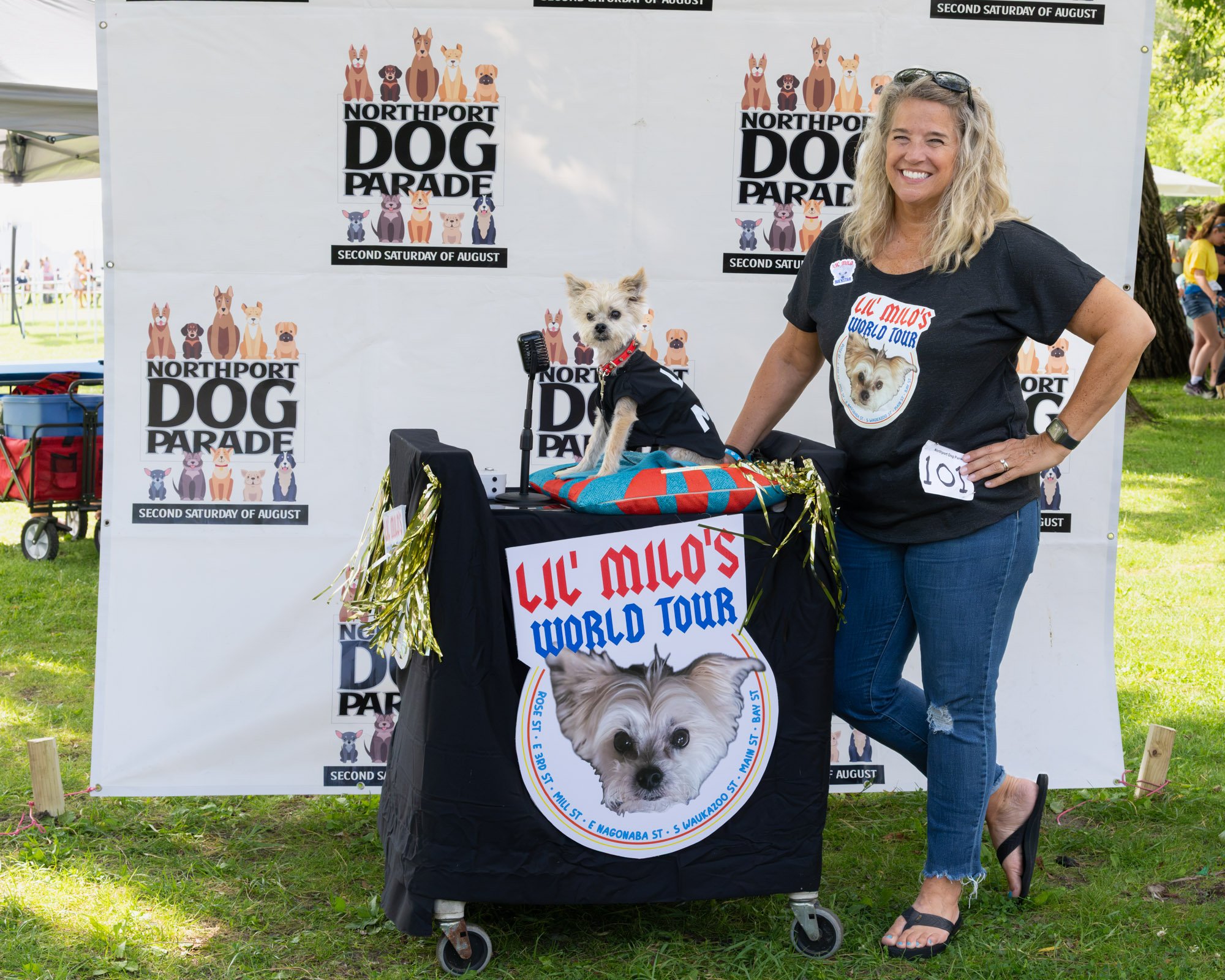 A woman with blonde hair and sunglasses on her head wearing a black t-shirt and ripped jeans standing at a pet parade event with a small dog on a mobility scooter. The backdrop has multiple posters for the Northport Dog Parade held on the second Satu