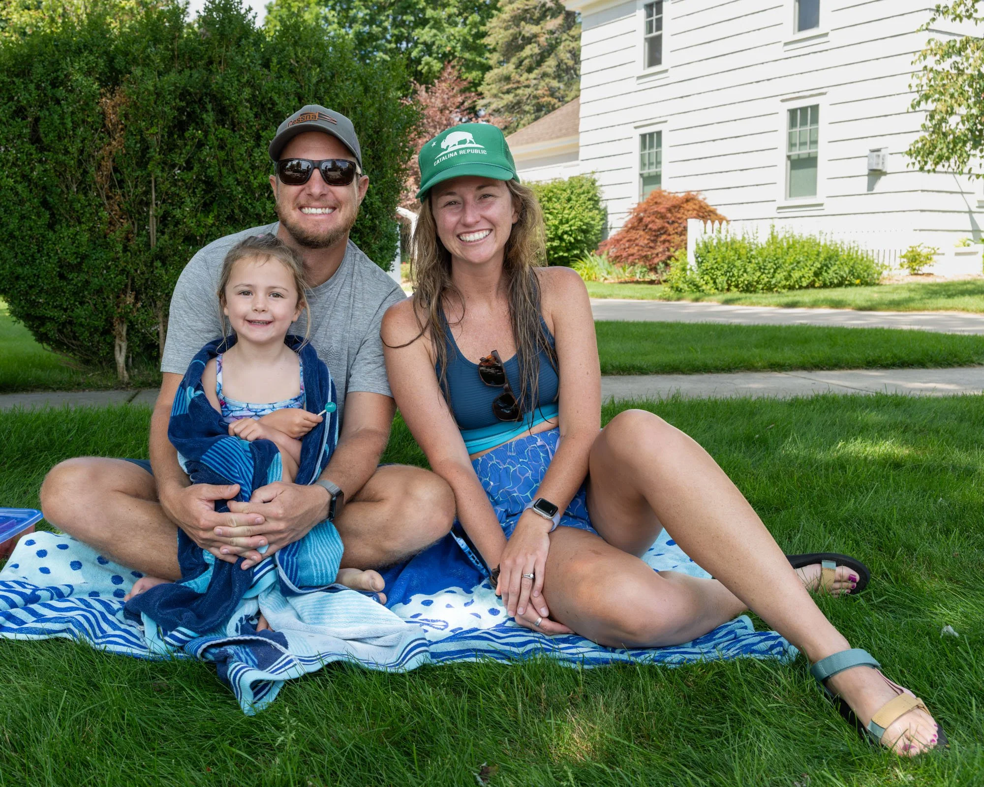A family of three sitting on a blue and white polka dot blanket on a grassy lawn in front of a white house with siding and multiple windows. The man is wearing sunglasses, a gray t-shirt, a gray cap, and has a beard. The woman is smiling, wearing a g
