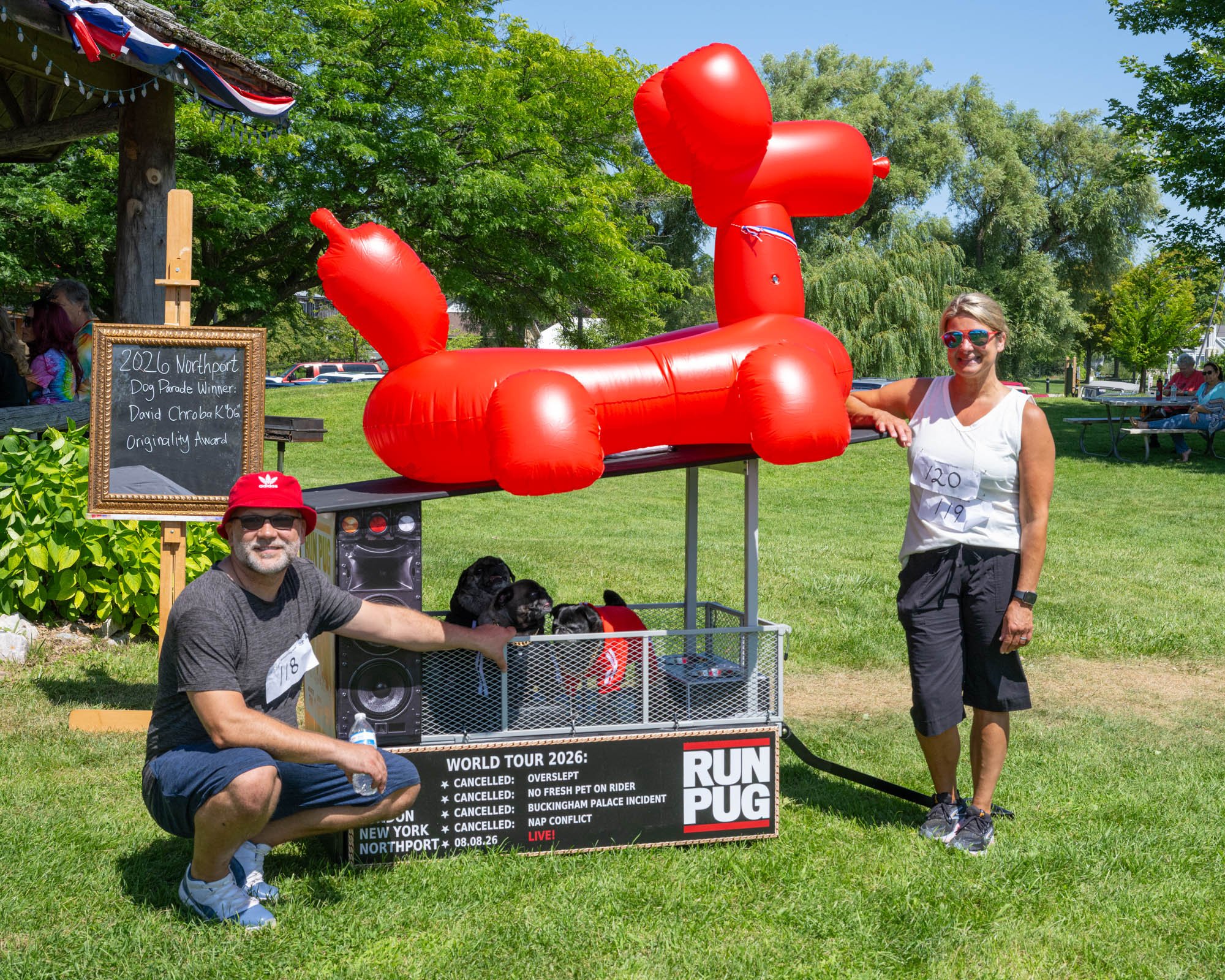 People standing around a red inflatable dog shaped float at an outdoor event in a park. One person is kneeling with a water bottle, and a woman is standing next to the float. There's a chalkboard sign with event details and a small black dog sitting 