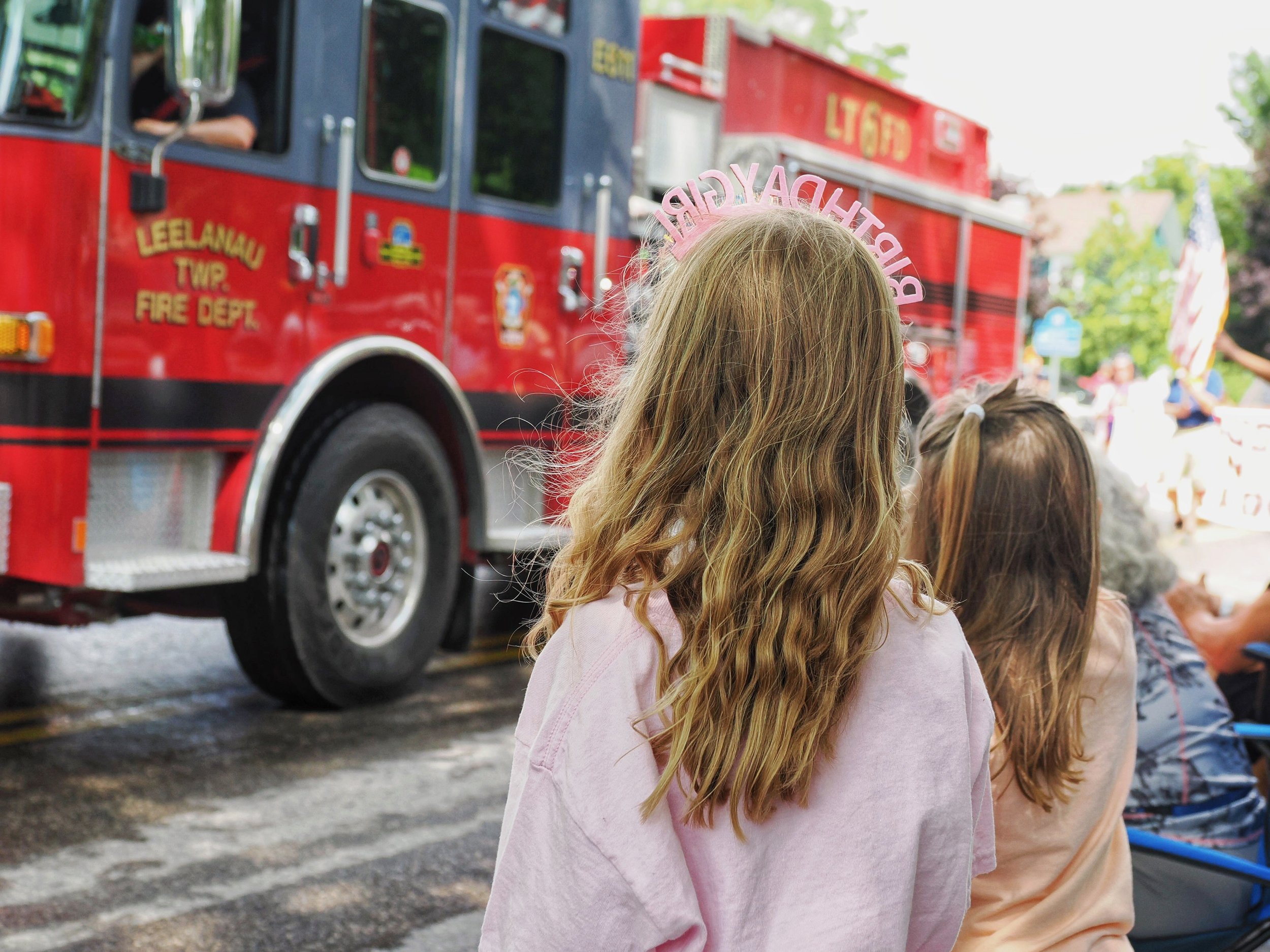Children sitting in line watching a fire truck during a parade or public event, with a girl in the foreground wearing a pink 'Happy Birthday' headband.