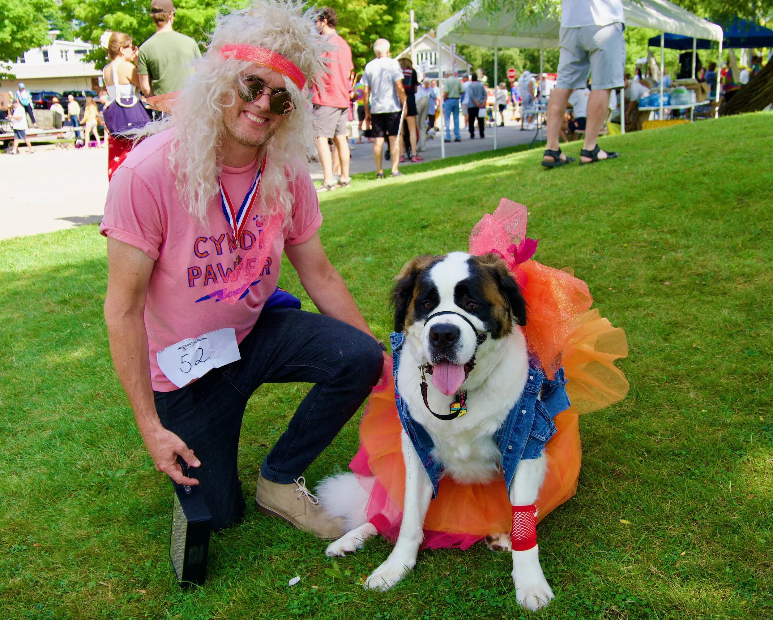 A man with long curly blond hair wearing a pink T-shirt with purple lettering, sunglasses, a red headband, and a medal around his neck is kneeling on grass next to a large dog dressed in colorful tutu and accessories. The dog appears to be a Saint Be