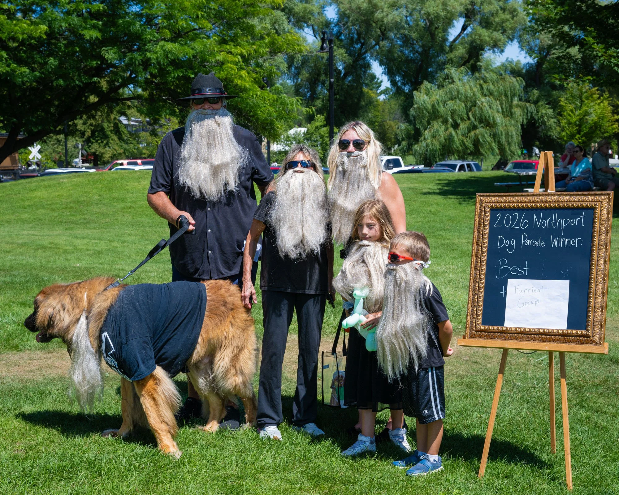 People dressed as bearded men, likely for a contest or parade, standing outdoors on grass with trees and cars in the background. A dog is in the foreground, and a chalkboard sign reads "2026 Northport Dog Parade Winner, Best & Furriest Group."