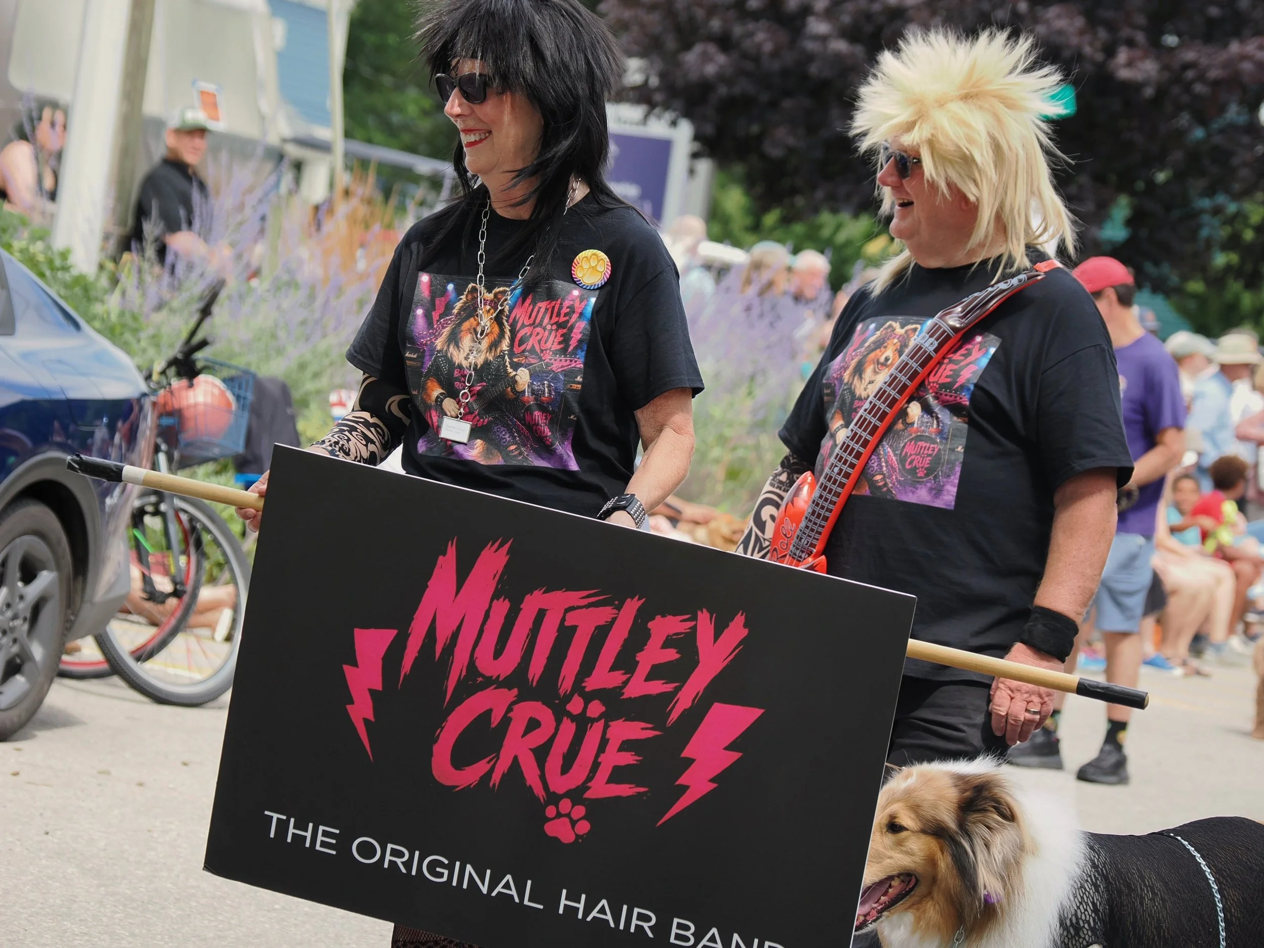 Two women with punk rock style hair and sunglasses at an outdoor event, holding a black sign with pink and red text that reads 'Motley Crue' and other words, with a dog nearby.