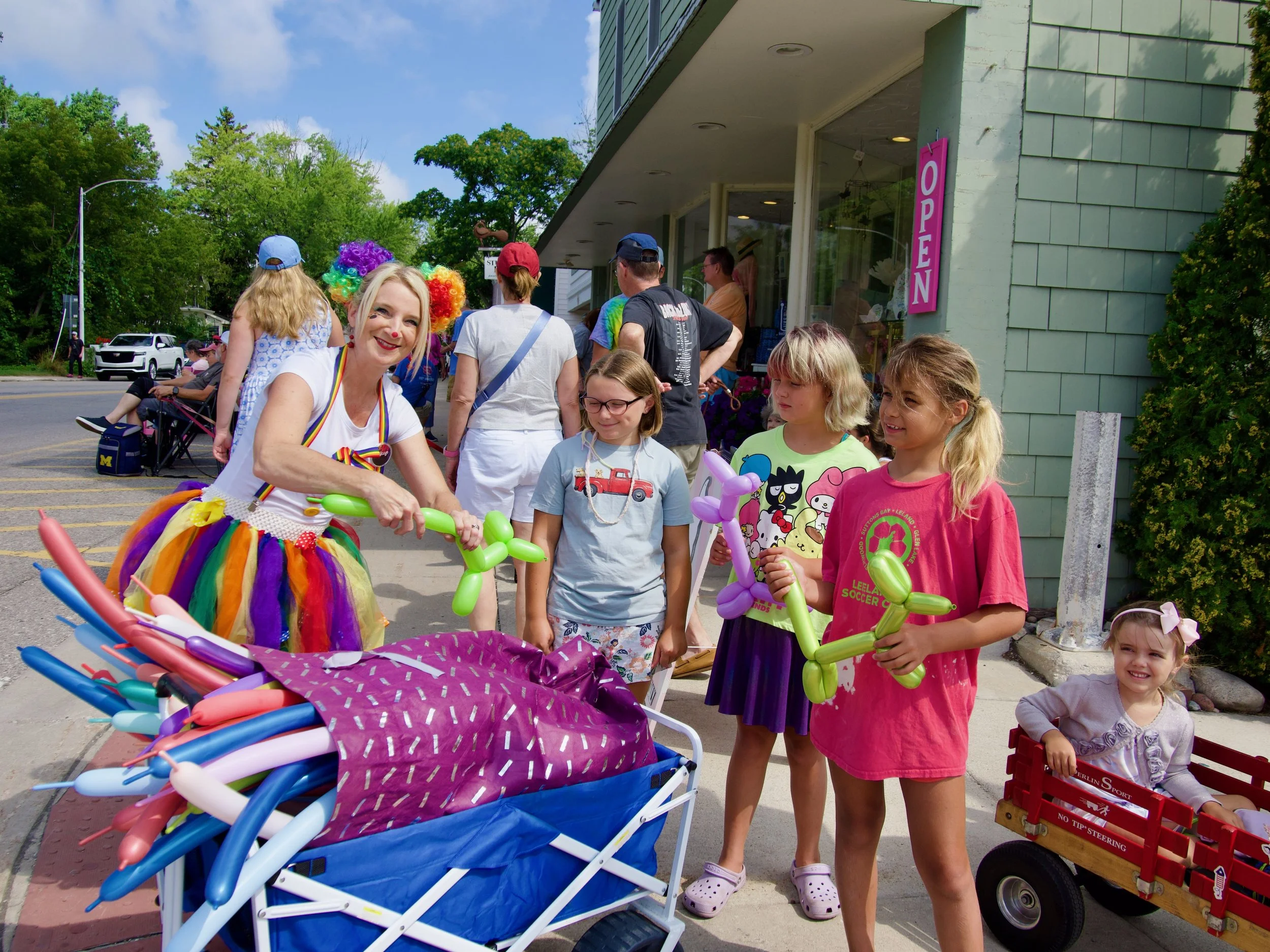 People celebrating an event outside a store with balloons, balloons shaped like animals, and colorful clothing, with trees and a sunny sky in the background.