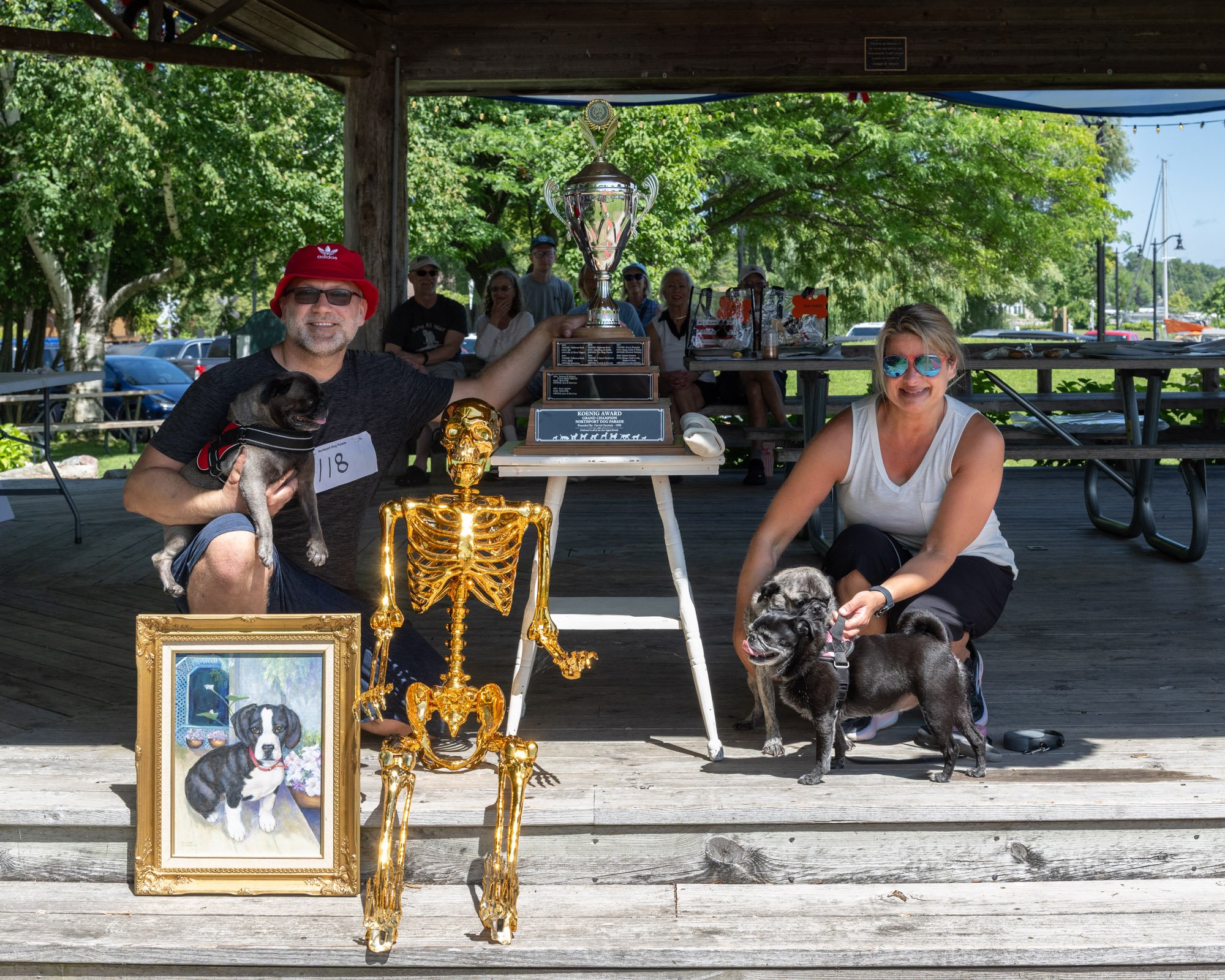 A group of people gathered outdoors under a pavilion, celebrating with trophies and dog portraits. A man is holding a small dog, and two large black dogs are in front. A woman is smiling, and others are sitting in background. There are trophies, a go