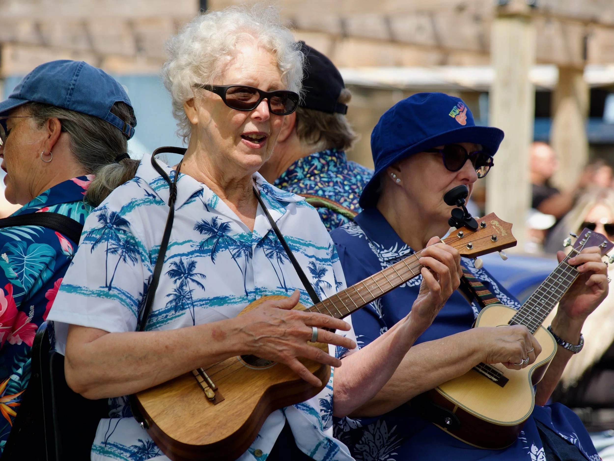 Group of older women outdoors, one playing a ukulele and another playing a guitar, wearing casual summer clothing and sunglasses.