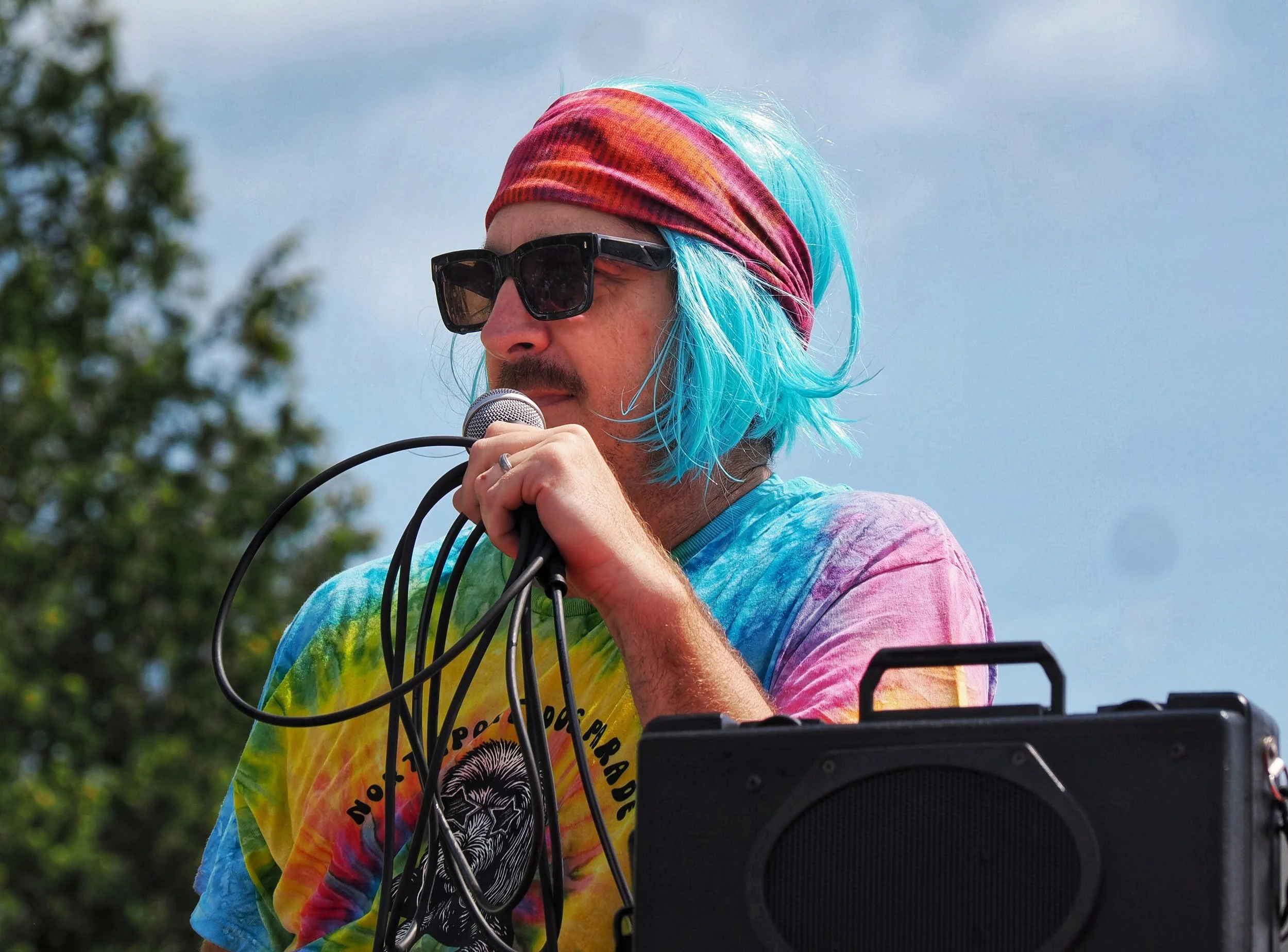 A person with blue hair wearing a colorful tie-dye shirt, a red headband, and large sunglasses, singing into a microphone outdoors with a blurry sky and some trees in the background.