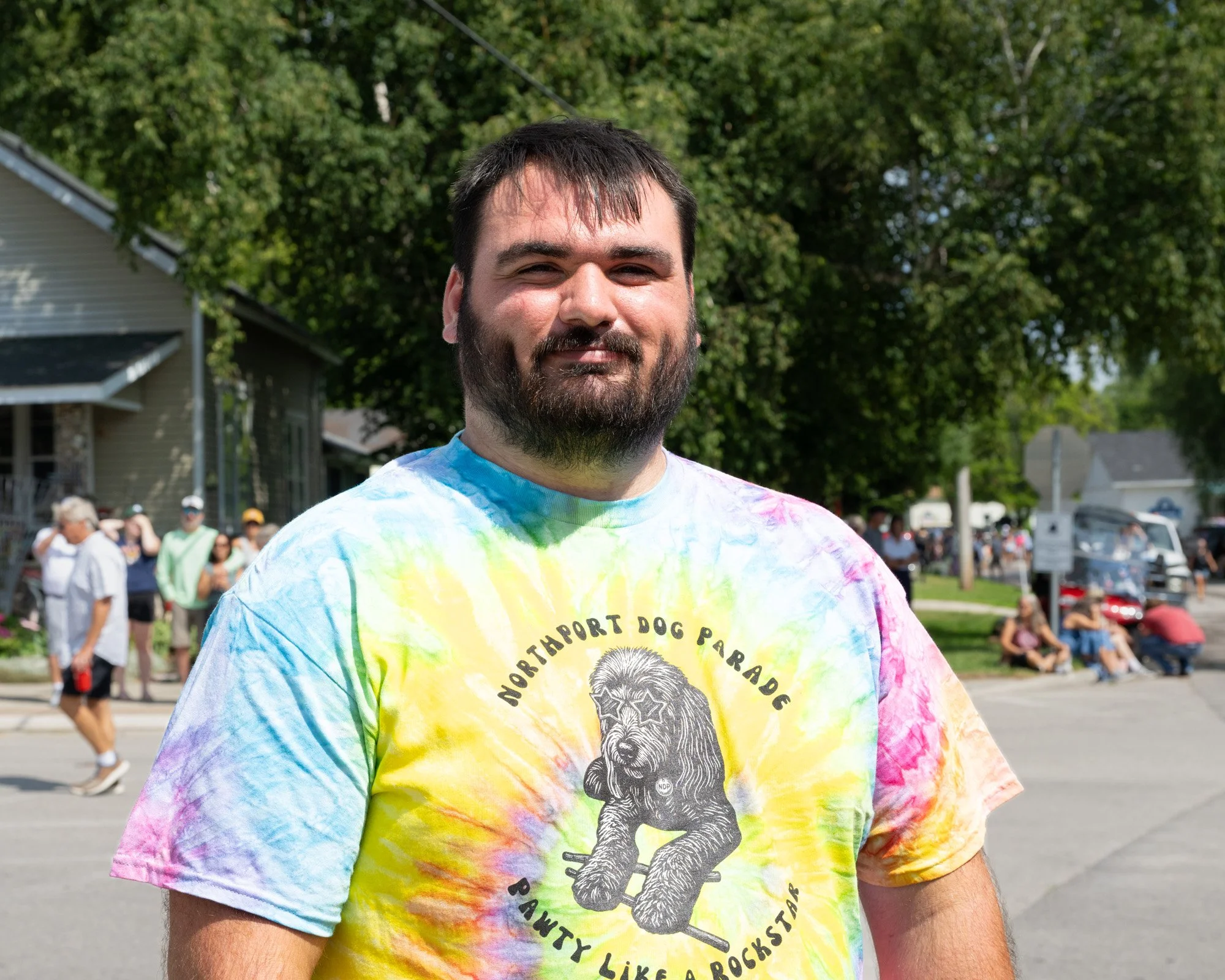 A man with dark hair and a beard smiling outdoors during daylight, wearing a colorful tie-dye T-shirt with a bear image and text, with a crowd and trees in the background.