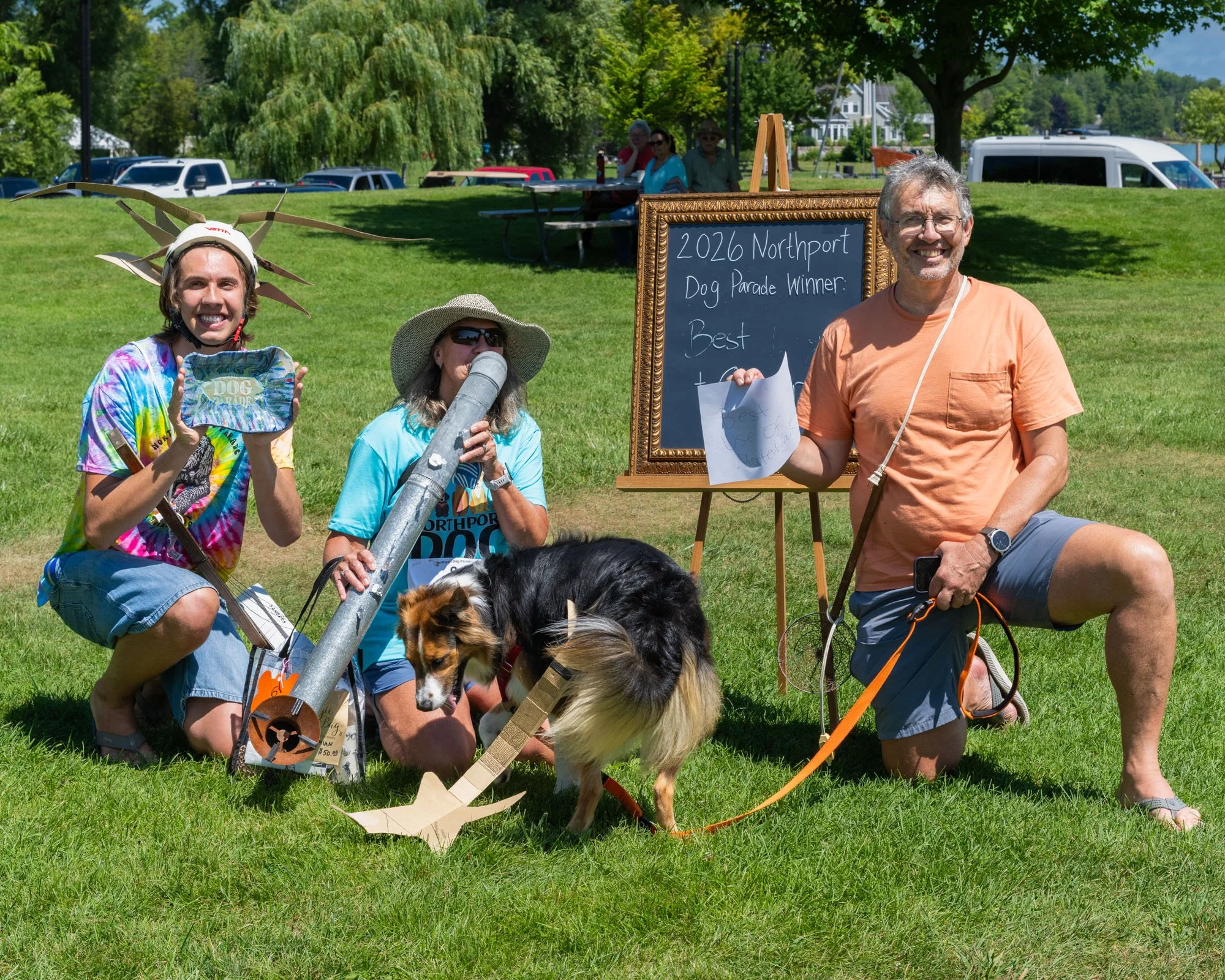 A group of people at a dog parade event in a park. One person is holding a dog with a costume resembling a chipmunk, and another person is drinking from a large silver container. There's a man holding a piece of paper next to a blackboard sign that r