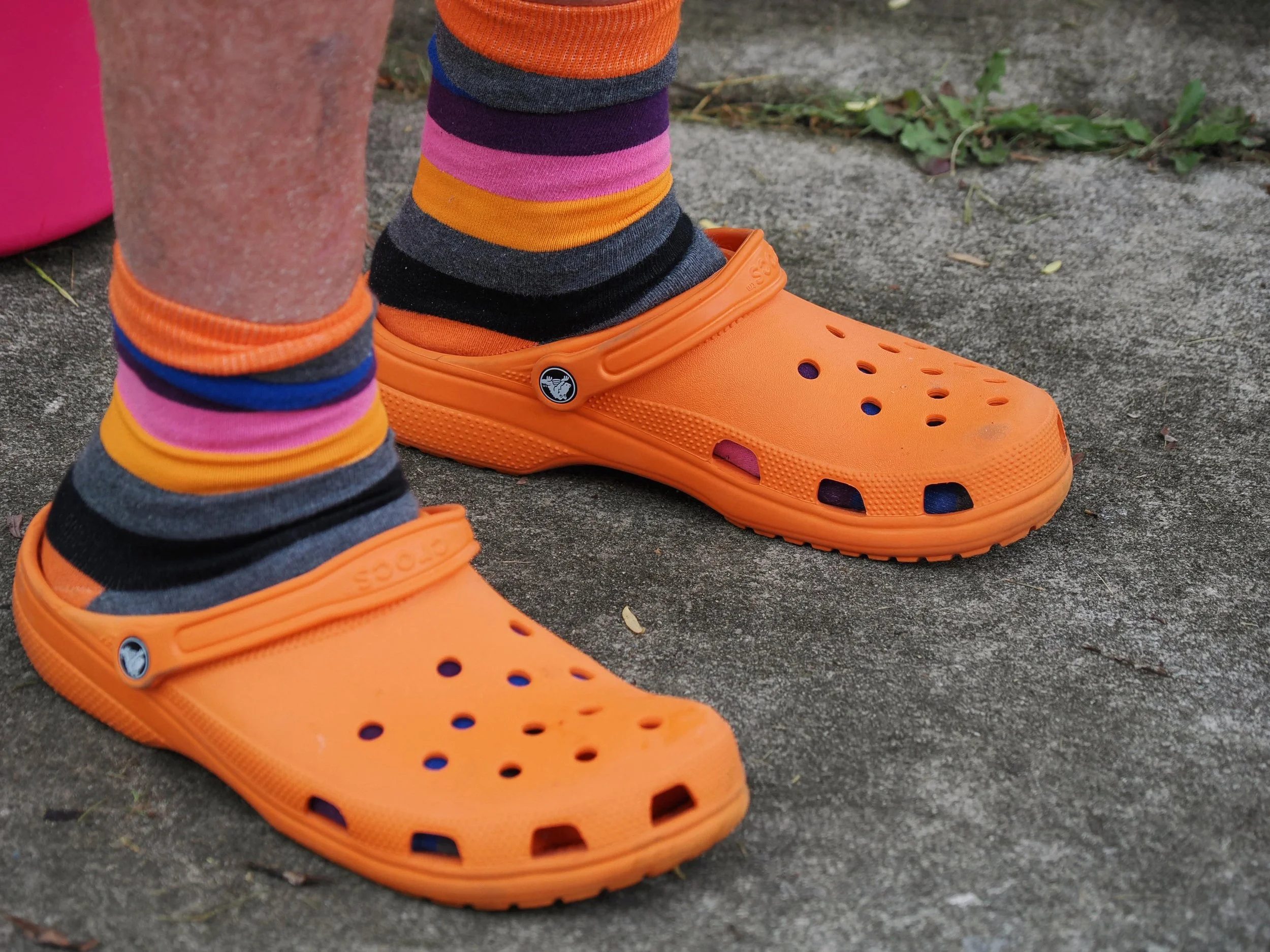 Close-up of a child's feet wearing orange Crocs with adjustable straps, striped colorful socks, standing on a concrete surface outdoors with small plants nearby.