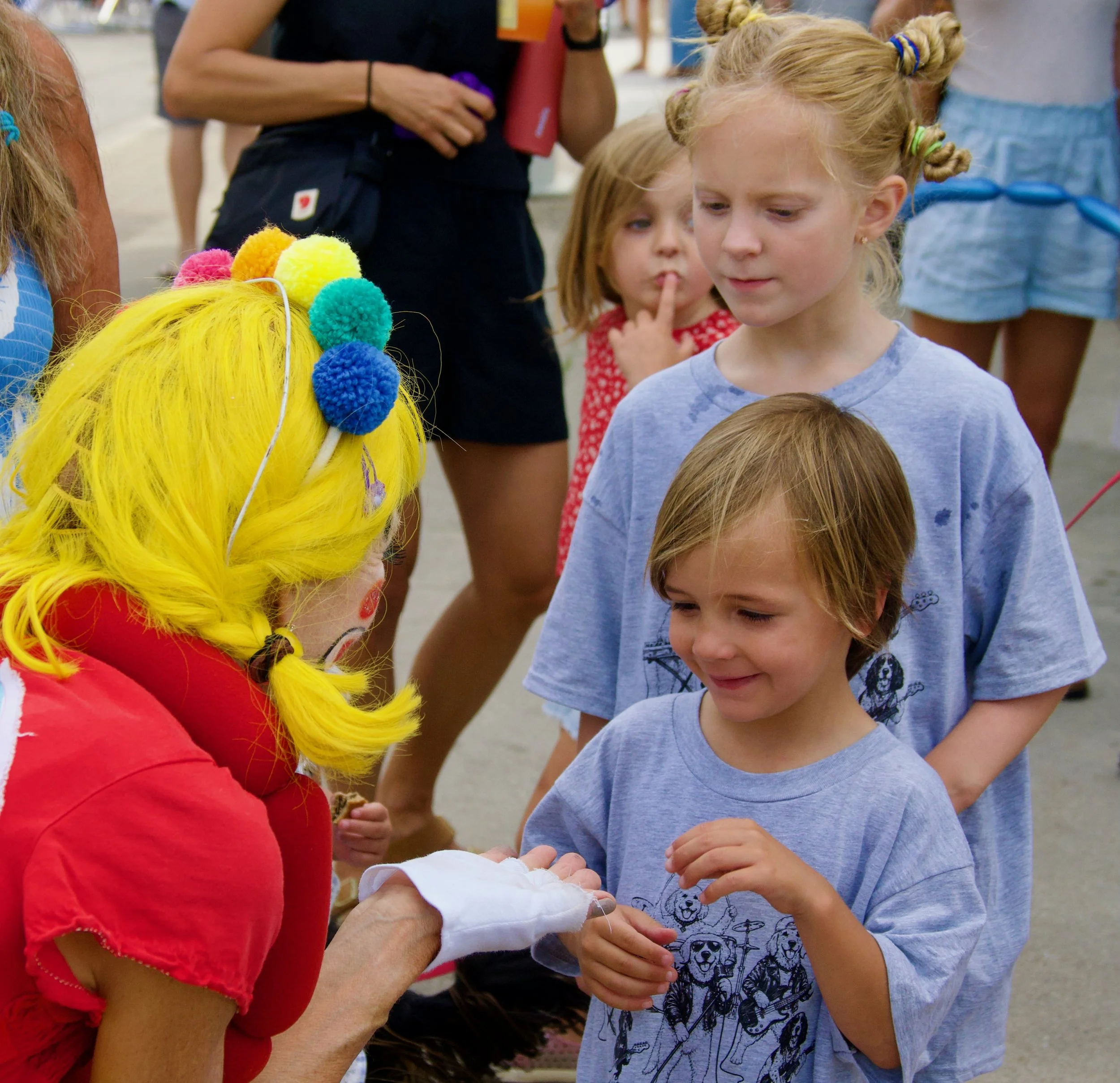Children gathered around a clown with a yellow wig decorated with colorful pompoms, engaging in an activity at an outdoor event.