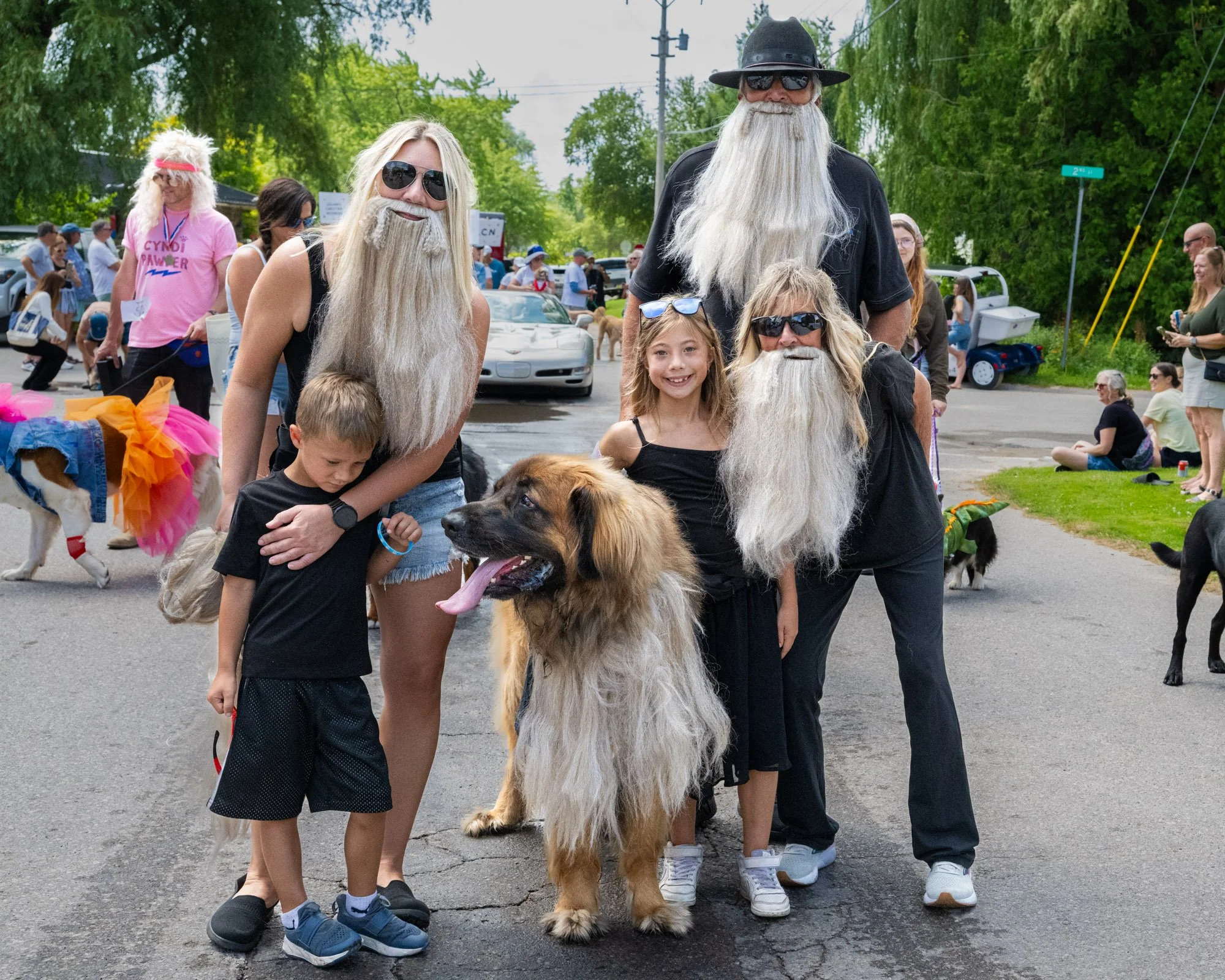 A group of people, including women, children, and large dogs, gathered outdoors during a sunny day, smiling and posing for a photo with two men dressed as Santa Claus with very long white beards and black hats, at an event in a park or street with gr