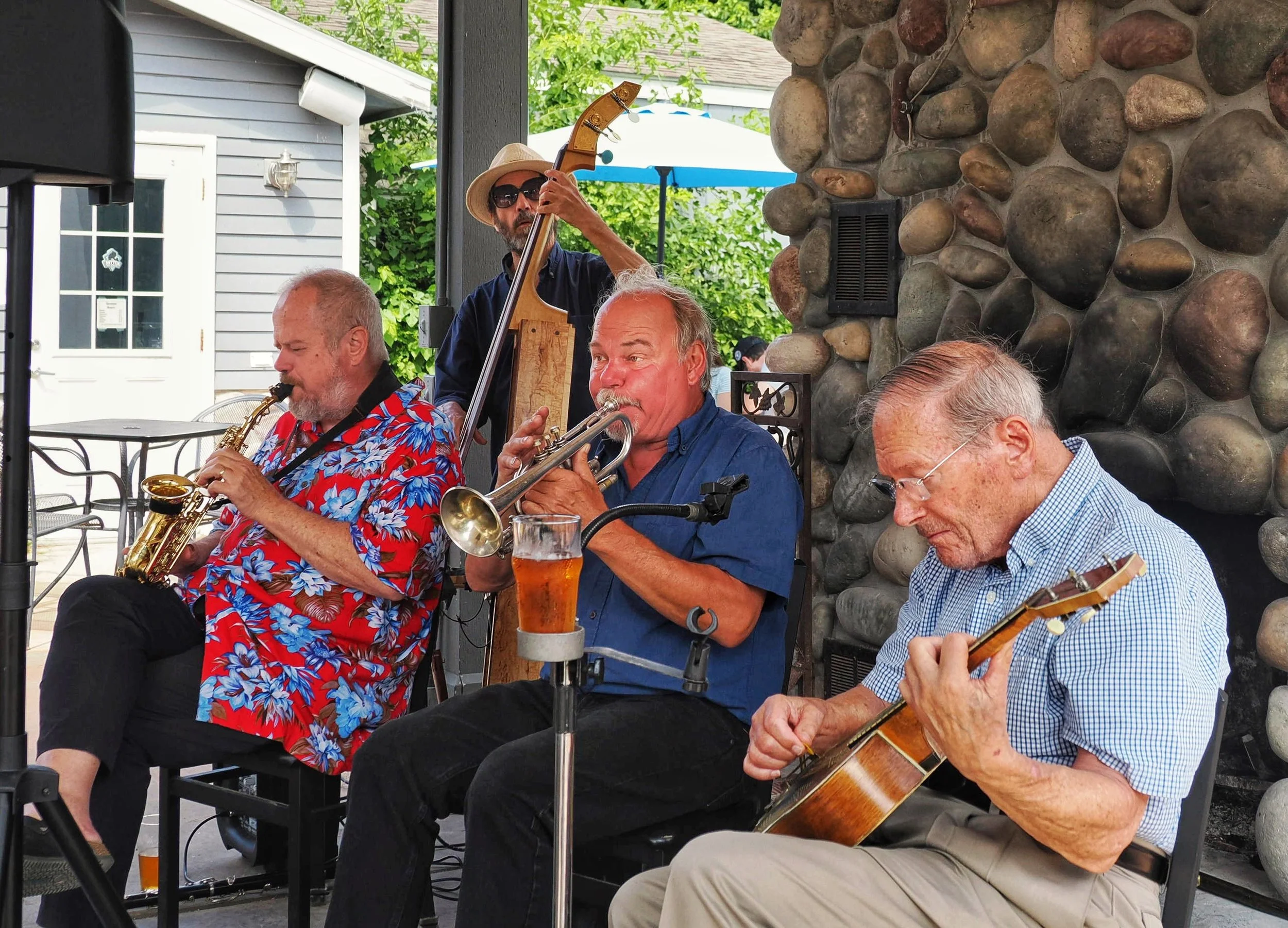 Four men playing musical instruments outdoors, with one man playing the saxophone, another the trumpet, and a third on guitar. There's a stone wall and greenery in the background, and a person with a hat playing the upright bass.