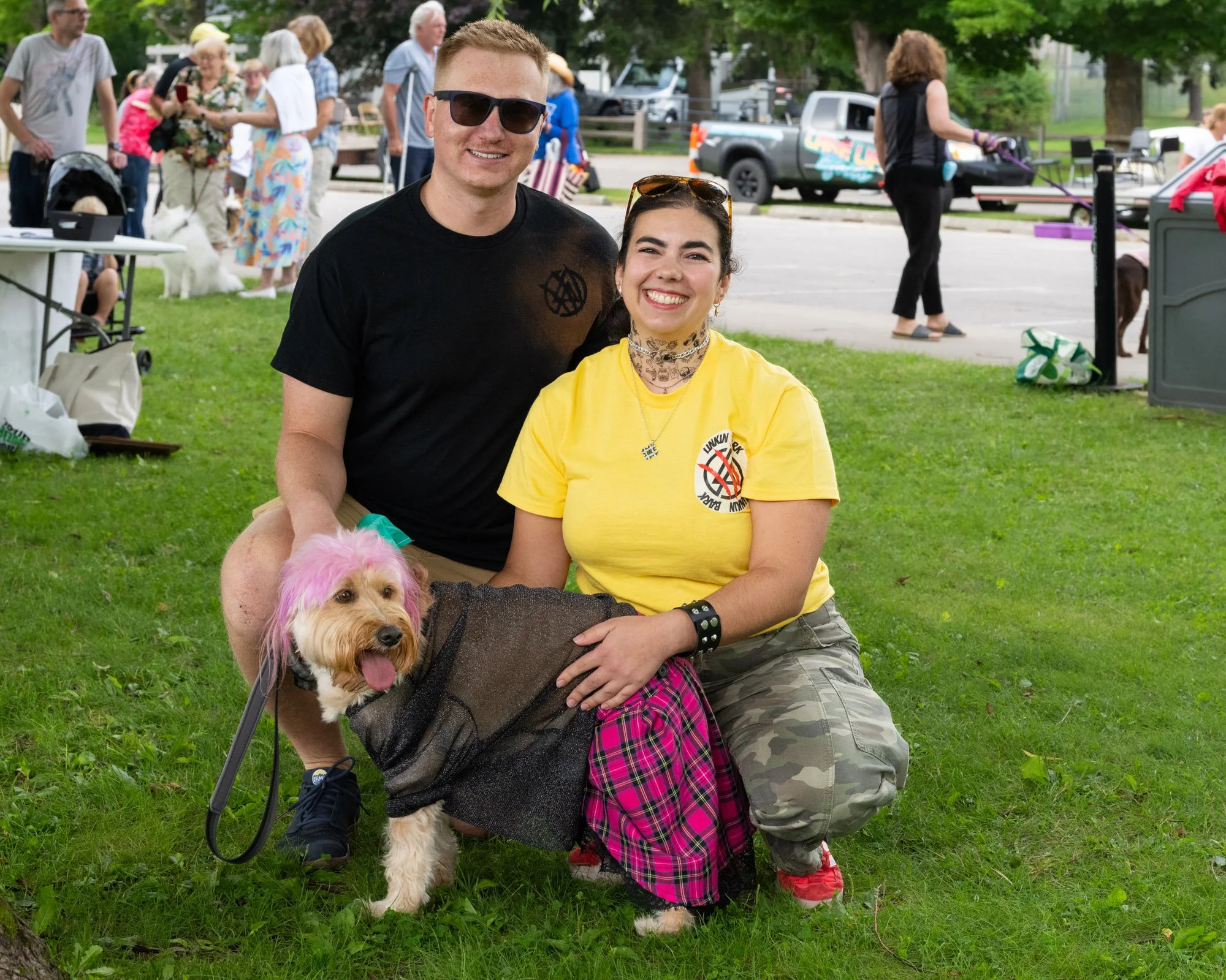 Two young adults, a man and a woman, smiling and posing outdoors at a park event, with a small dog that has pink hair on its head in front of them. The man is wearing sunglasses and a black t-shirt, and the woman is wearing a yellow t-shirt and patte