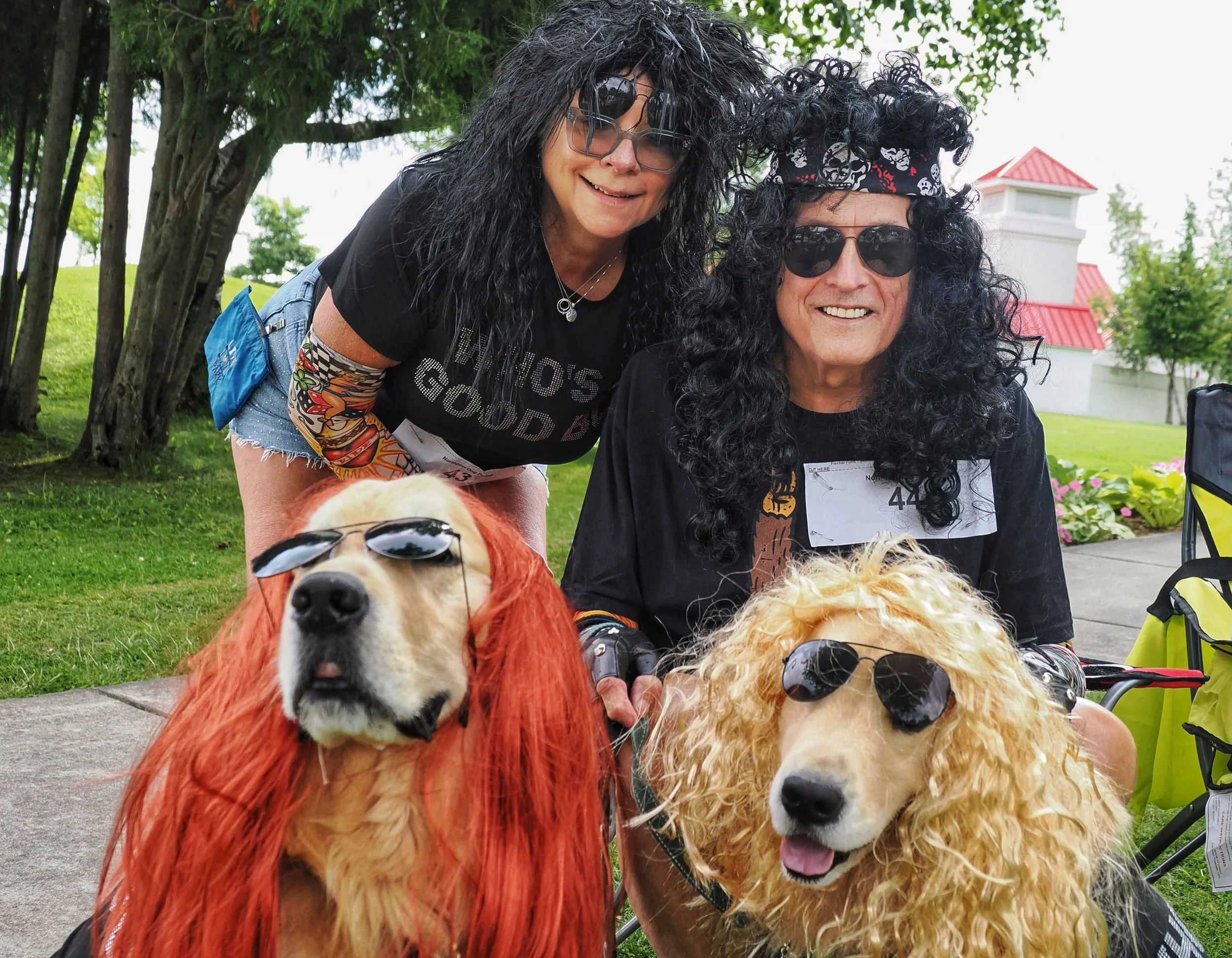 Two women dressed as rock stars with sunglasses and wigs, and two dogs with sunglasses, outdoors in a park with green trees and a building with red roofs in the background.