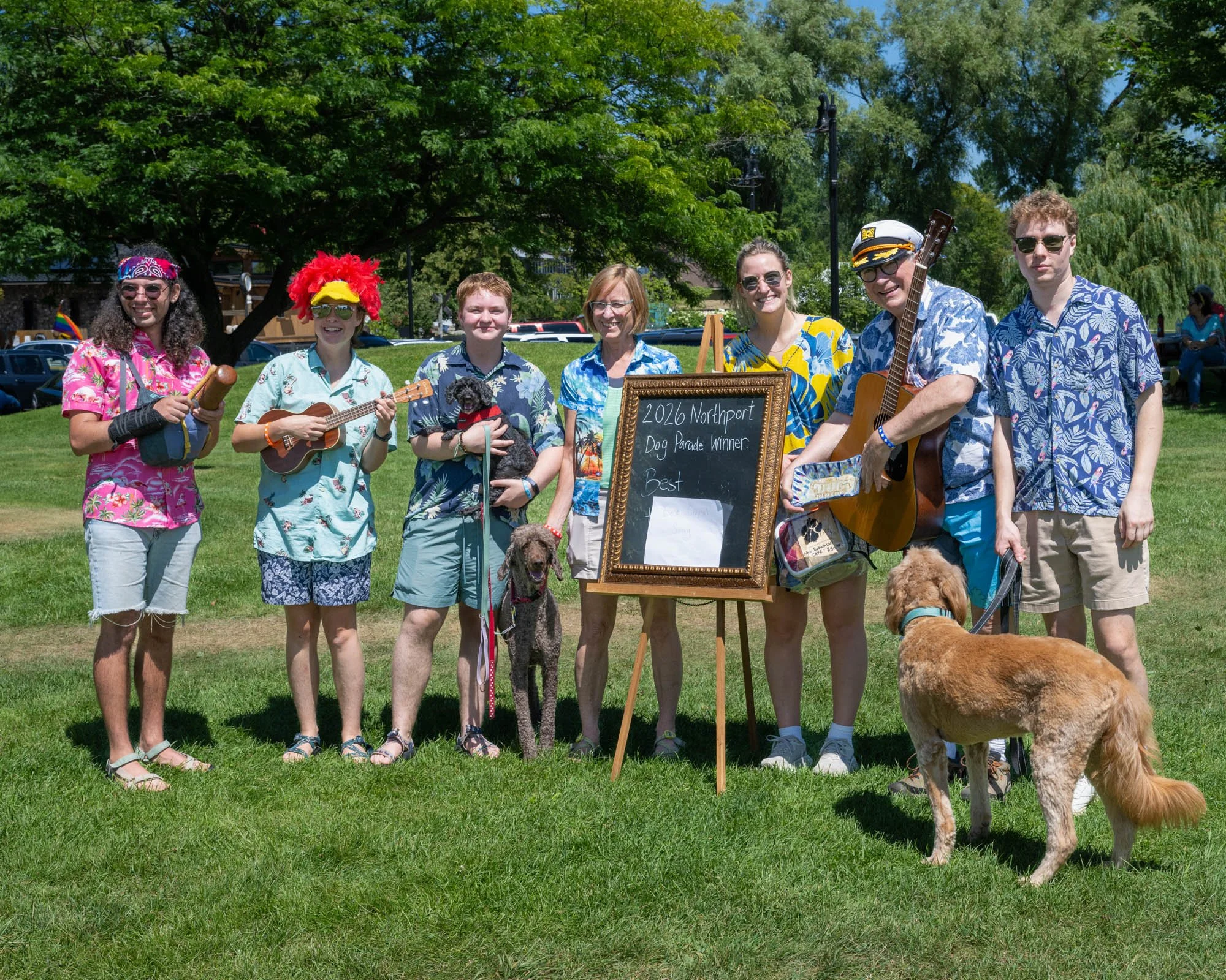 Group of seven people and three dogs standing outdoors on grass, with trees and parked cars in the background. Some people are dressed in colorful, tropical-themed clothing and holding musical instruments, with one person wearing a captain's hat and 