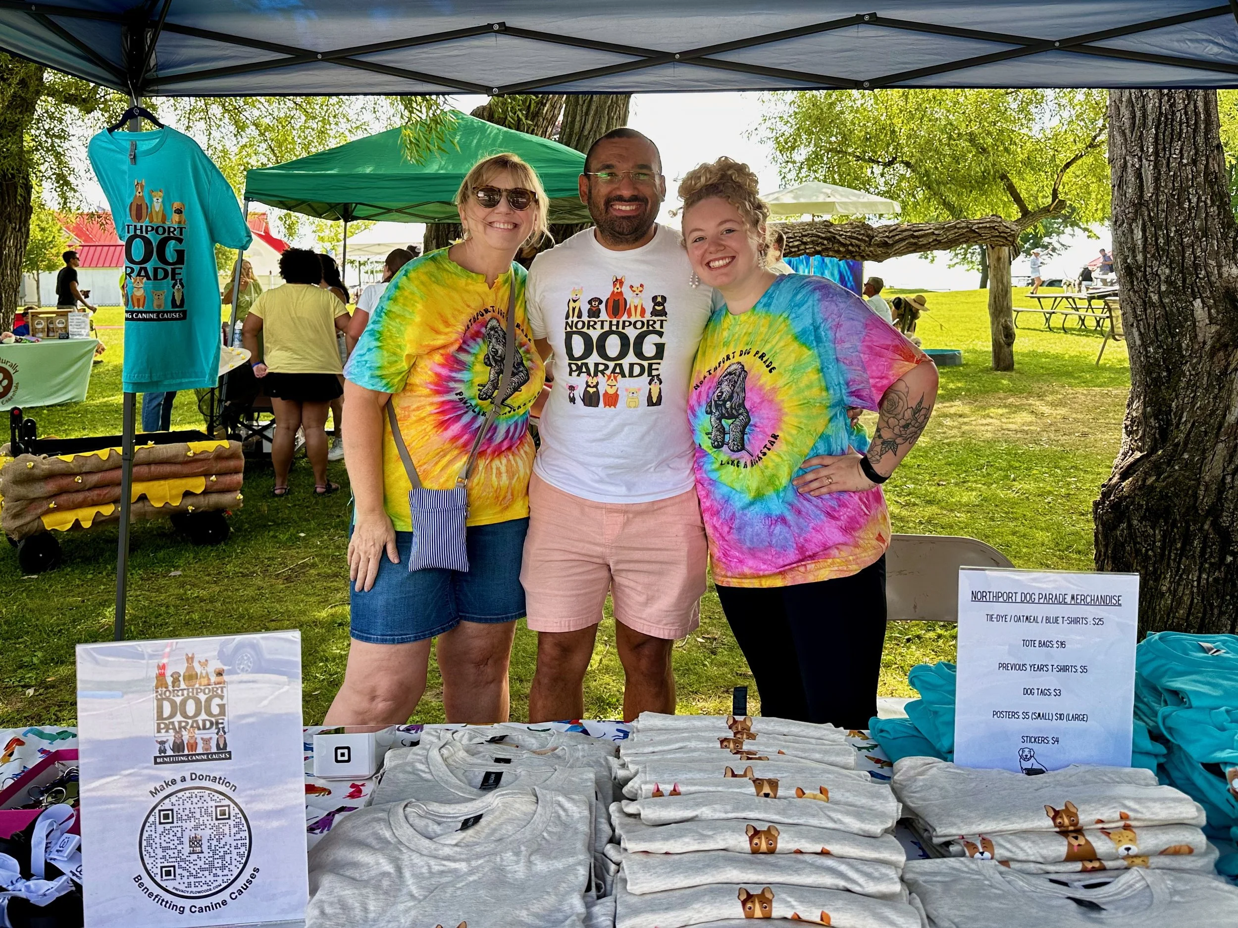 Three people smiling at a booth during an outdoor event, with merchandise including T-shirts and tote bags related to the Northport Dog Parade, under a canopy in a park.