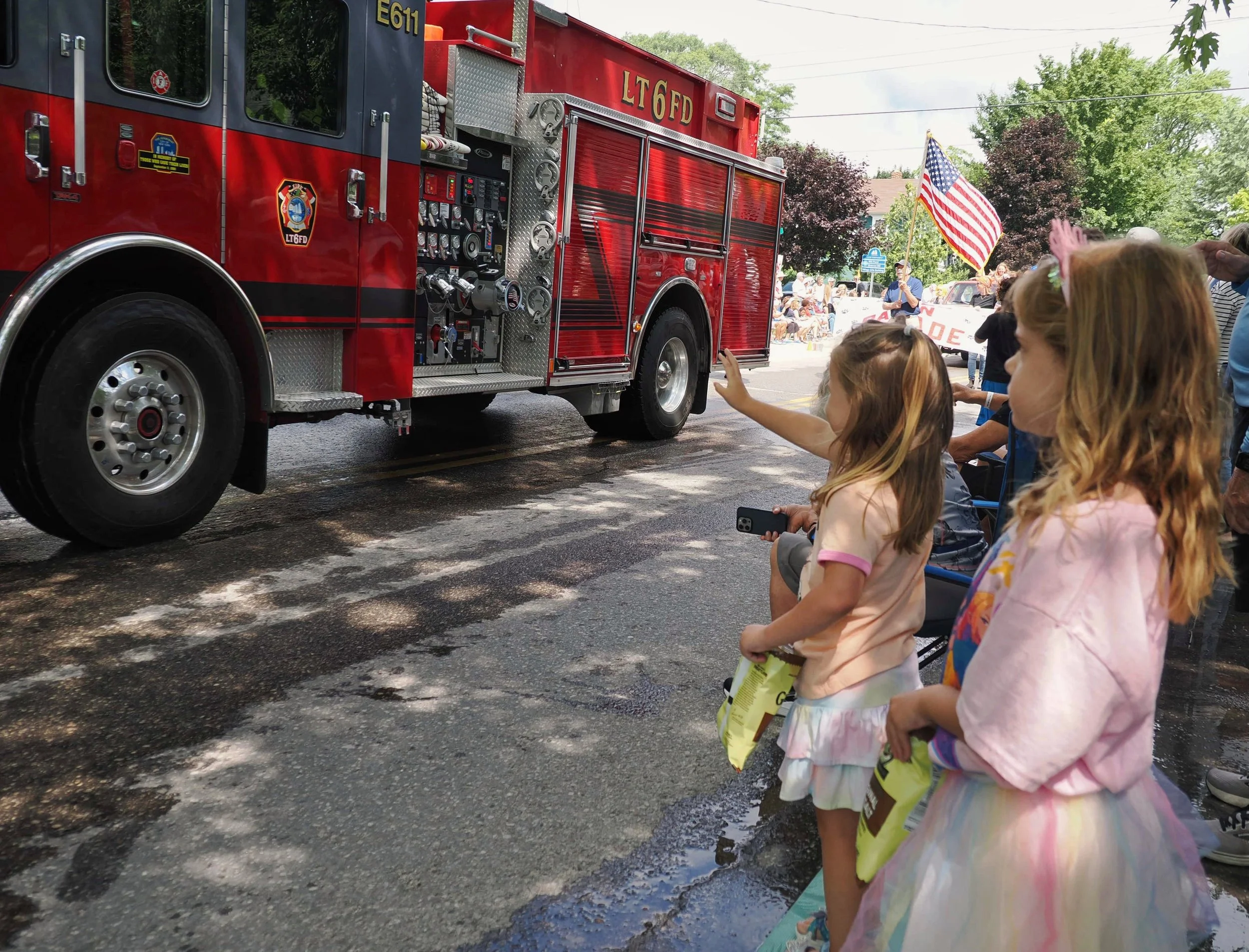 A red fire truck with children and adults gathered around during a parade or community event. An American flag is being held by a person in the background, and some children are reaching out towards the fire truck.
