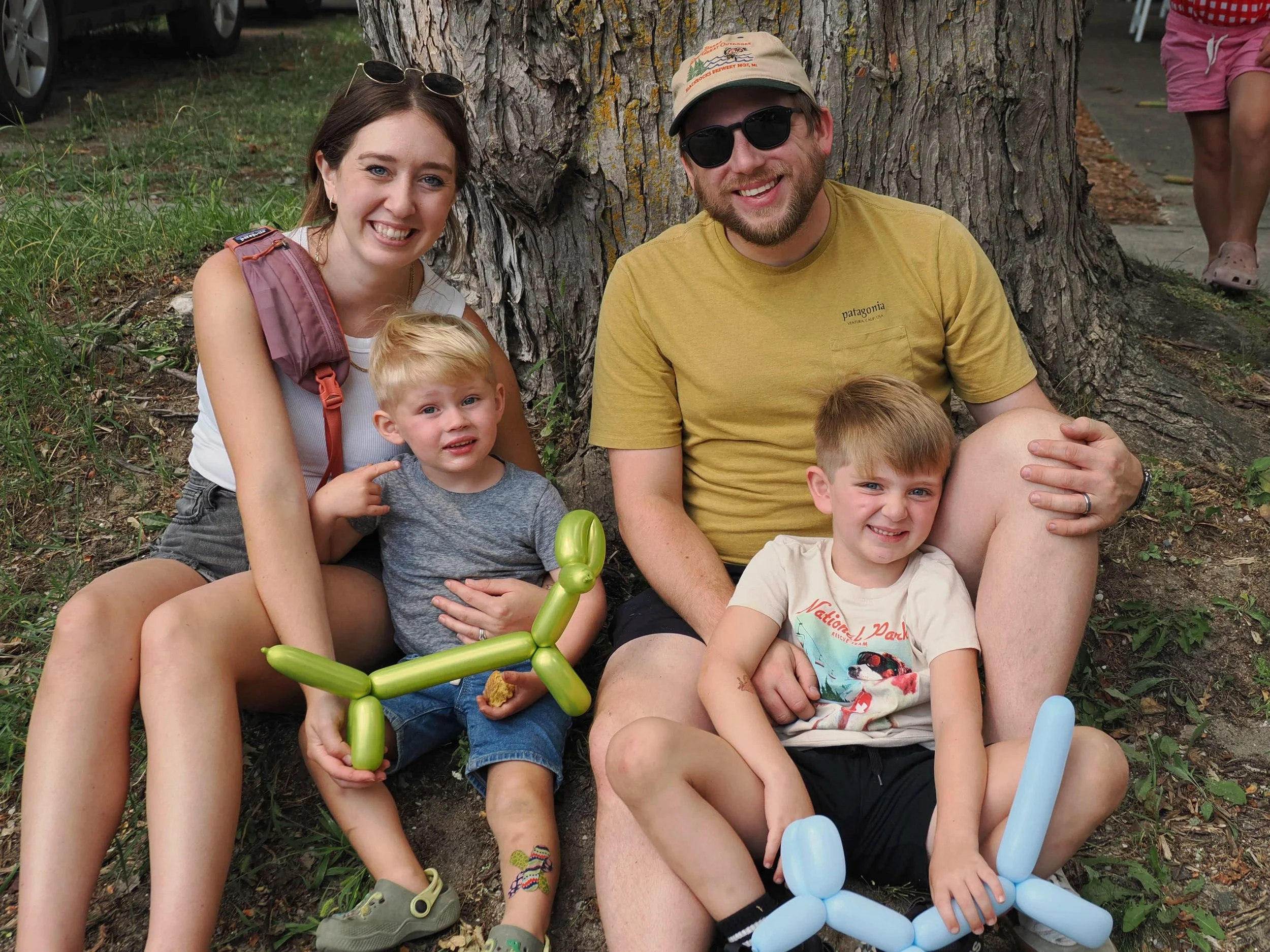 A smiling family of four sitting on the grass in front of a large tree, with a woman, a man, and two young boys, one of whom is holding a green balloon animal, and there are balloons on the ground.