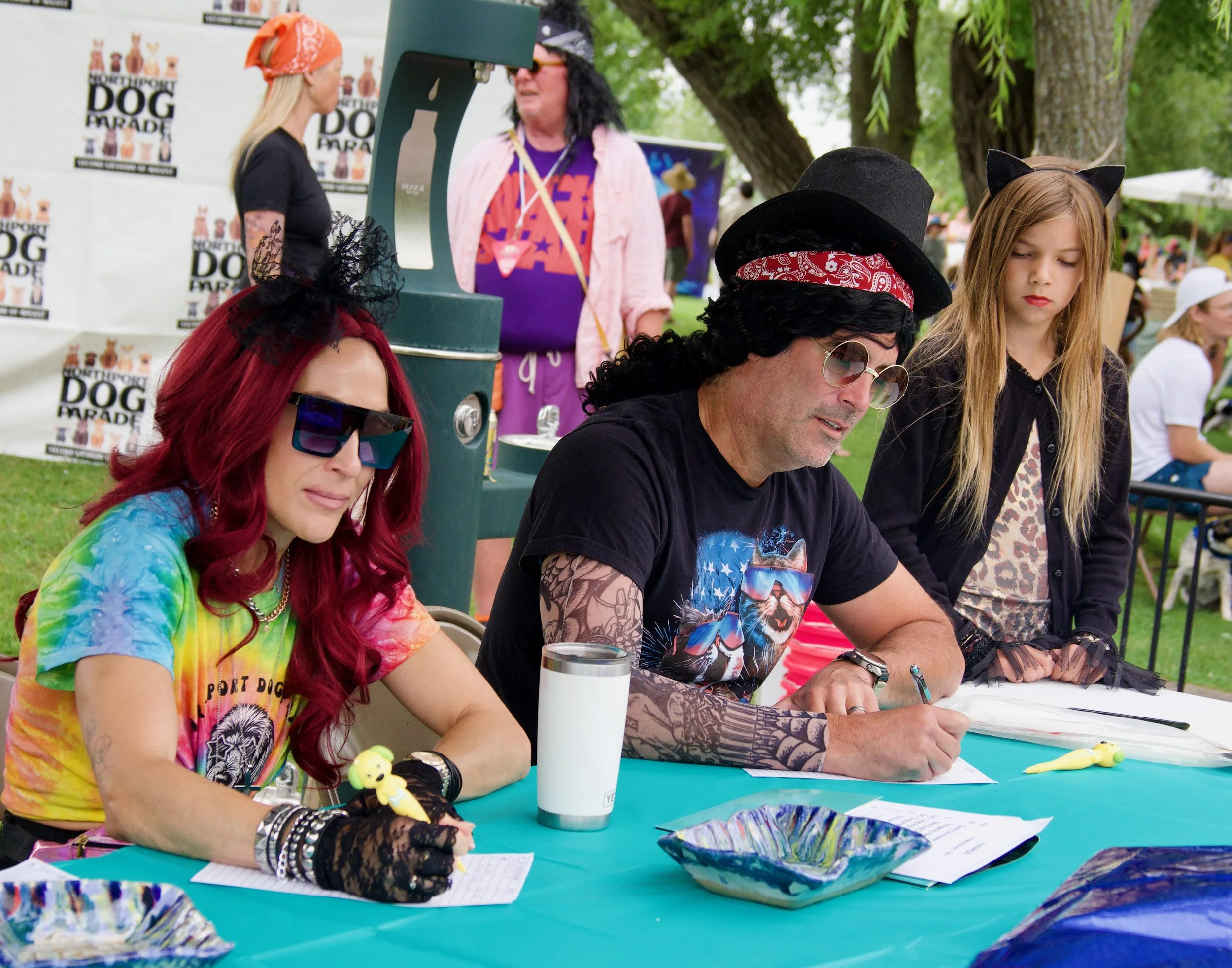 People dressed in colorful and themed costumes sitting at a table outdoors at a festival, with some wearing cat ears, bandanas, and sunglasses.