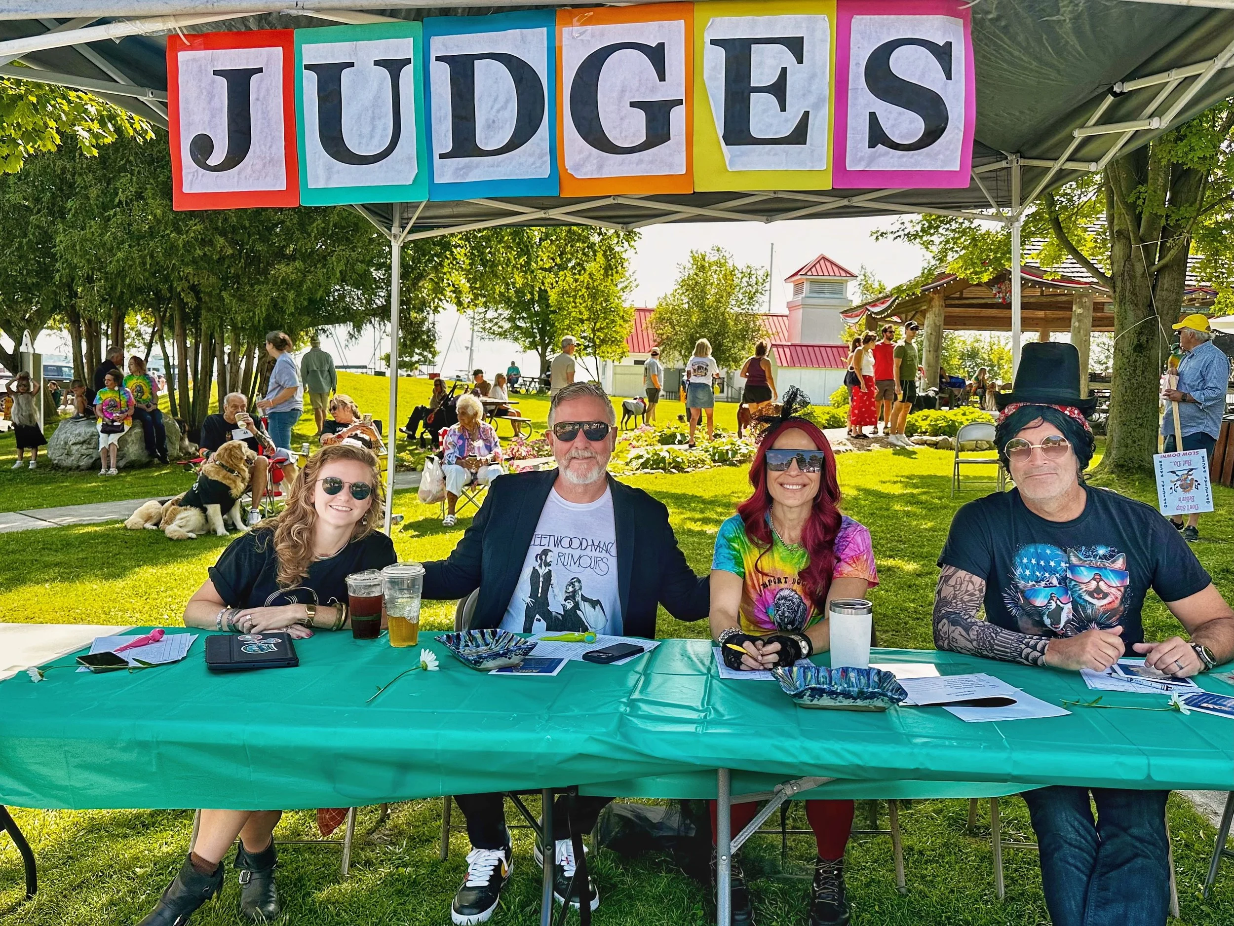 A group of people sitting and standing under a canopy at an outdoor event with colorful signs that spell 'JUDGES' hanging above. There are tables with papers and drinks in the foreground, and other attendees and greenery in the background.
