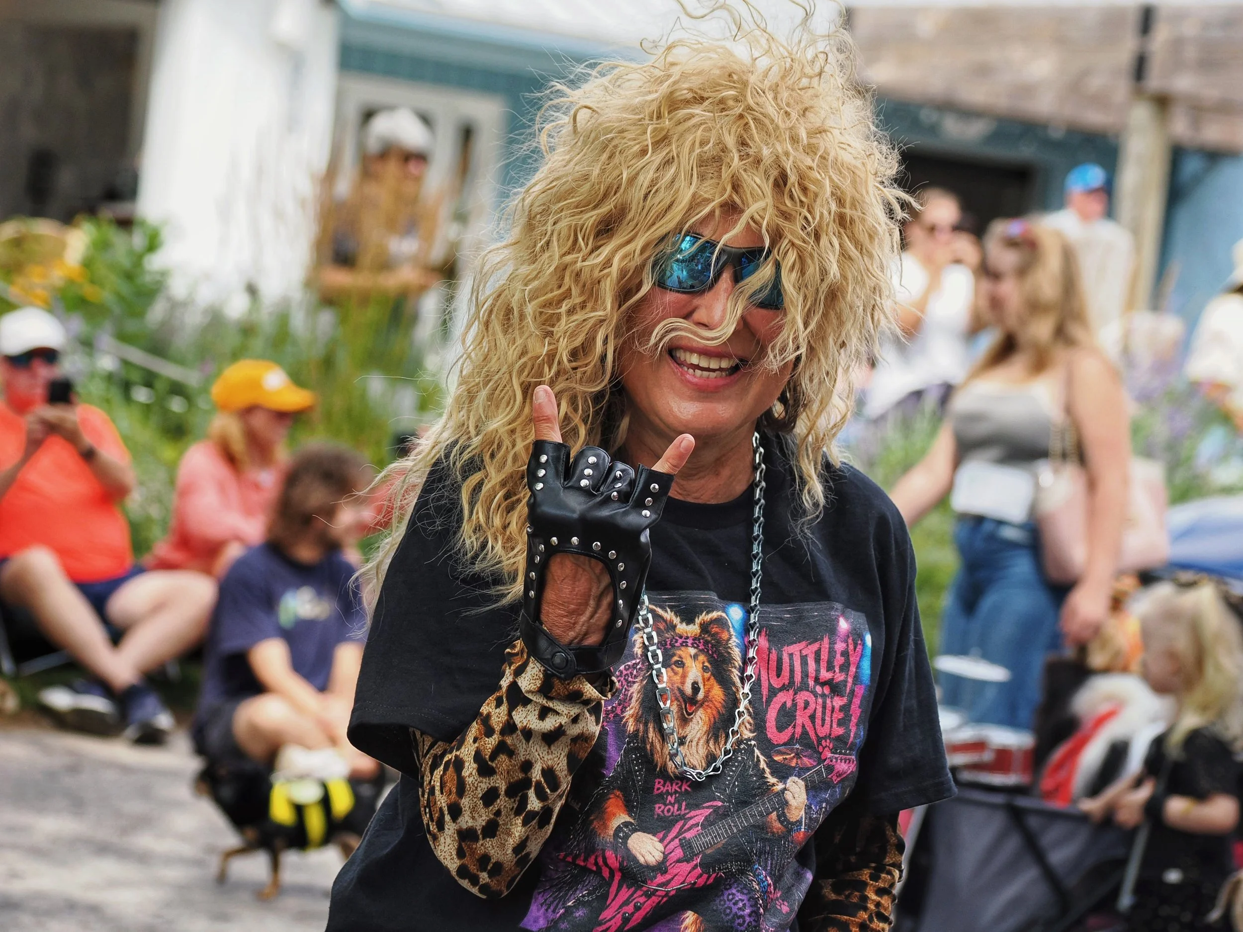 A woman with curly blonde hair wearing dark sunglasses and a black t-shirt with colorful graphics, including lions and guitars, is smiling and making a peace sign with her right hand. She is also dressed in a leopard print sleeve and a large chain ne