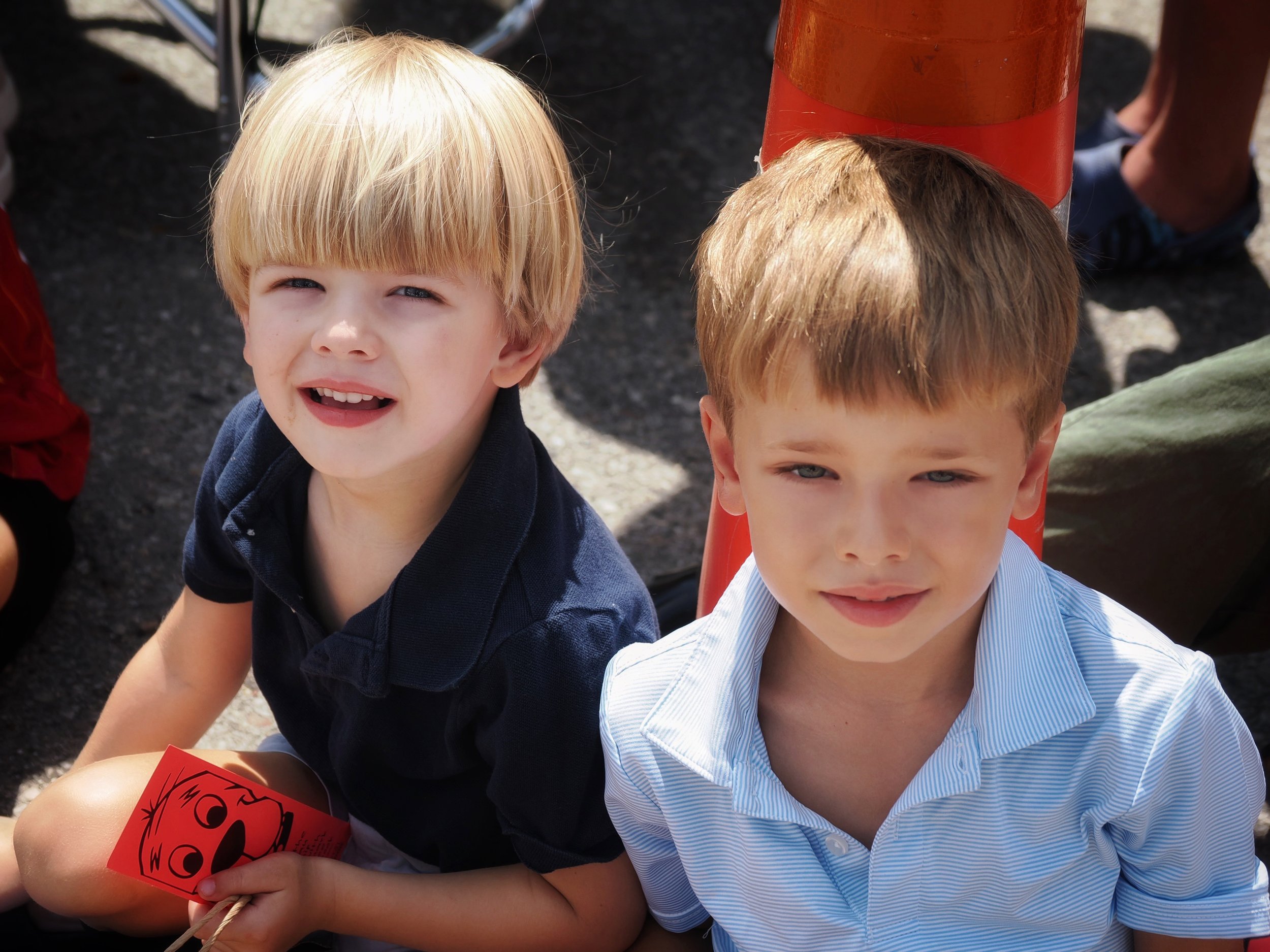Two young boys sitting outside; one with blond hair and one with light brown hair, both smiling and looking at the camera. The boy with blond hair is holding a small red paper with a black drawing on it.