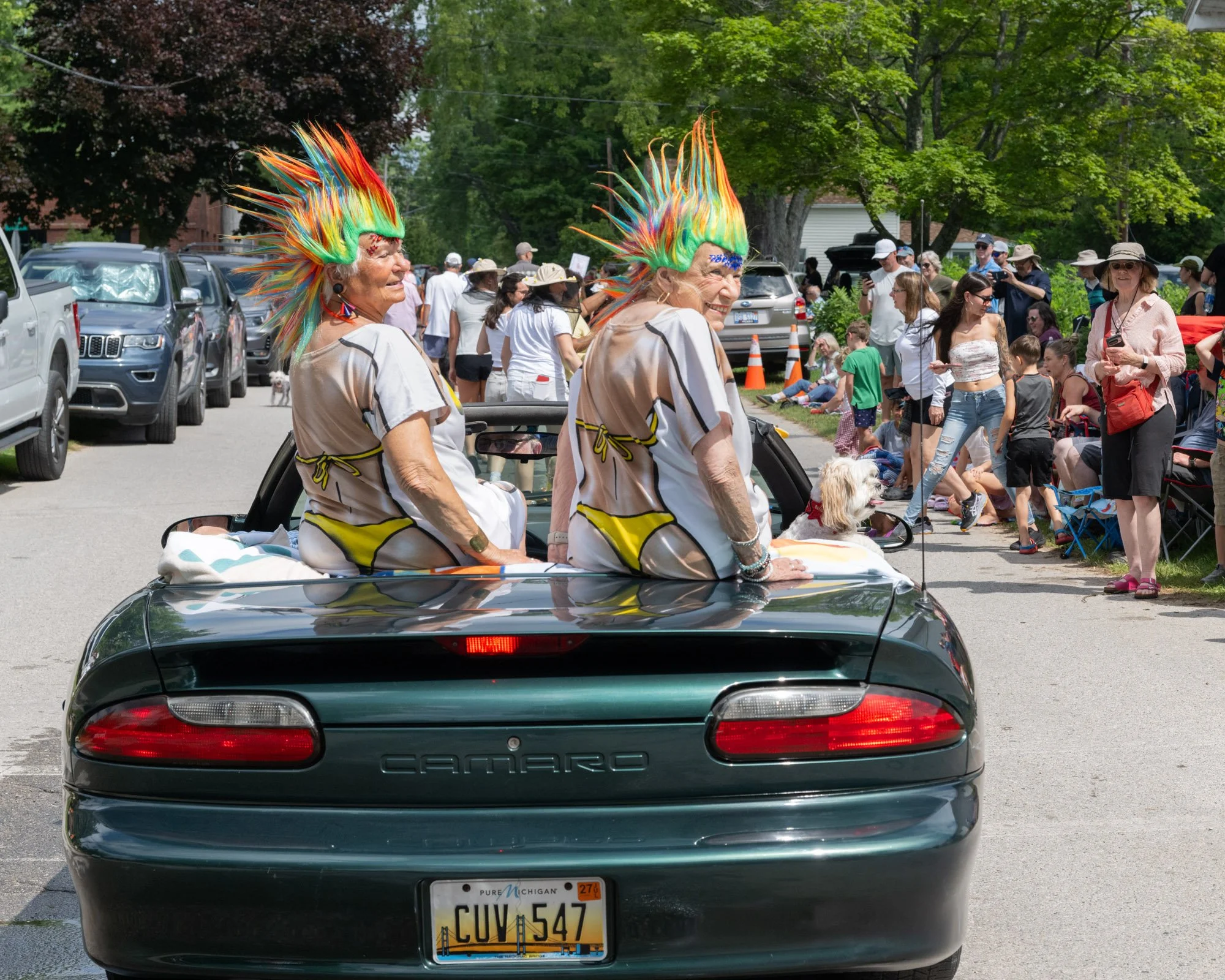 A black Chevrolet Camaro with a Michigan license plate. Two elderly women with colorful, spiked hair headbands are in the back seat, smiling and enjoying a parade or outdoor event on a sunny day. There are many onlookers and parked cars along the str