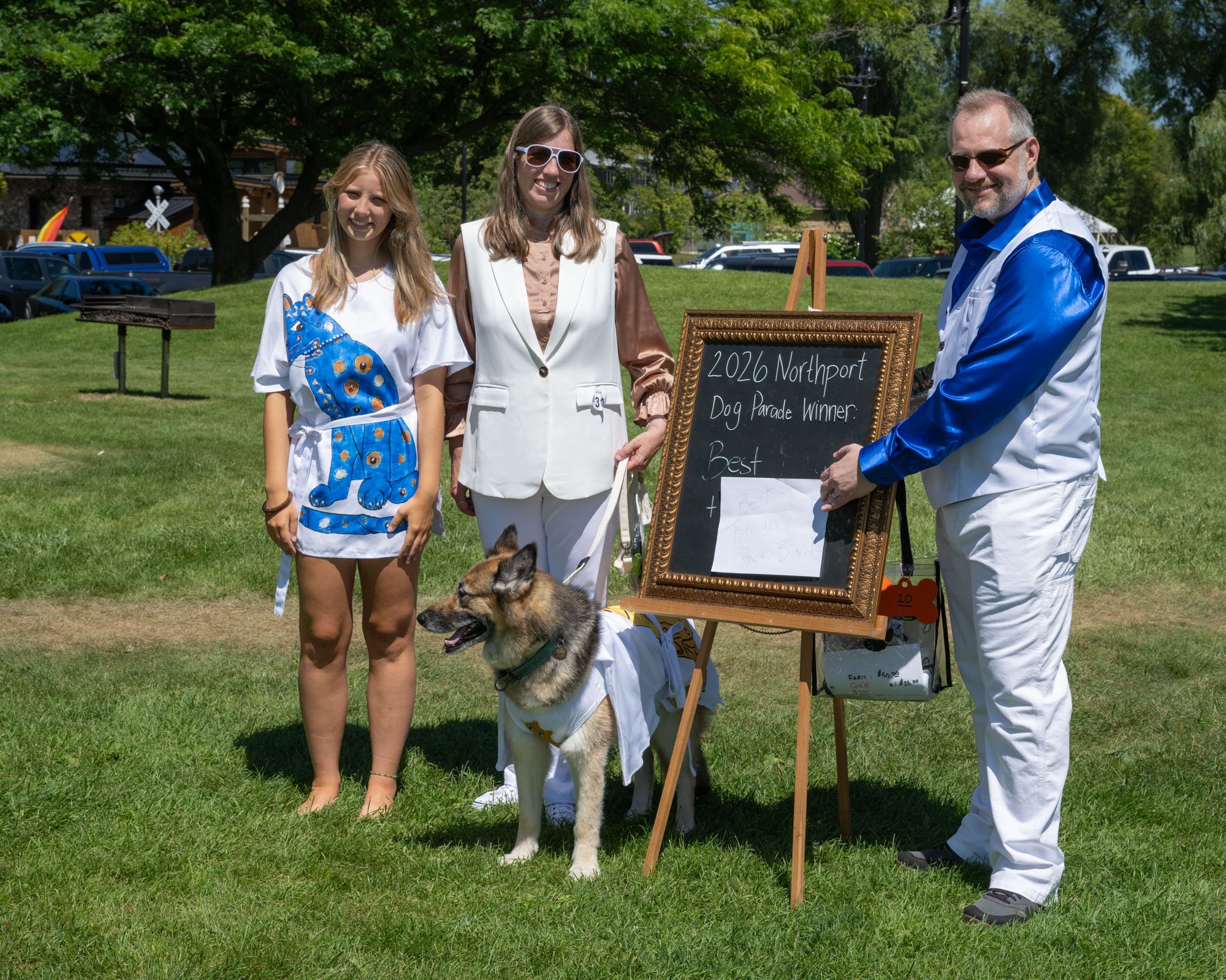 Three people and a dog outdoors during daytime at a park or event. The young girl on the left is smiling, wearing a white t-shirt with a blue dog graphic. The woman in the middle is smiling, dressed in a beige blazer with a white vest, and wearing wh