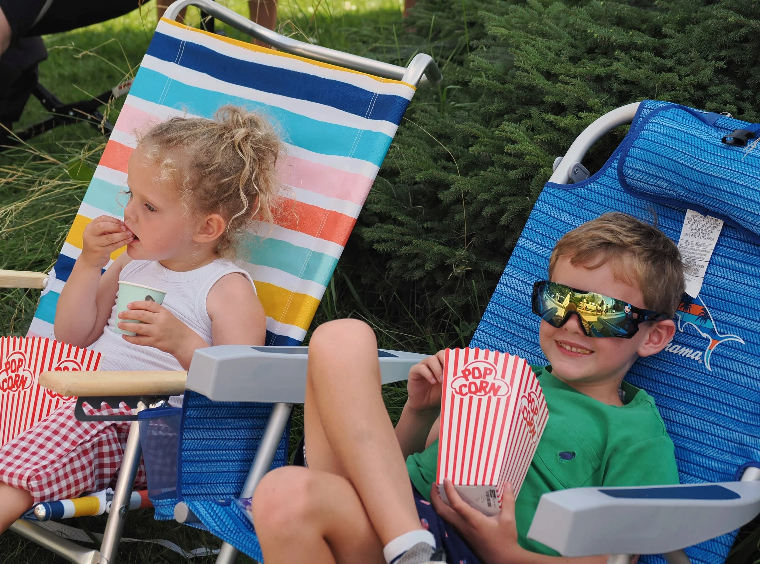 Two children sitting outdoors in reclining chairs, eating popcorn and enjoying snacks at a picnic or rest area near greenery.