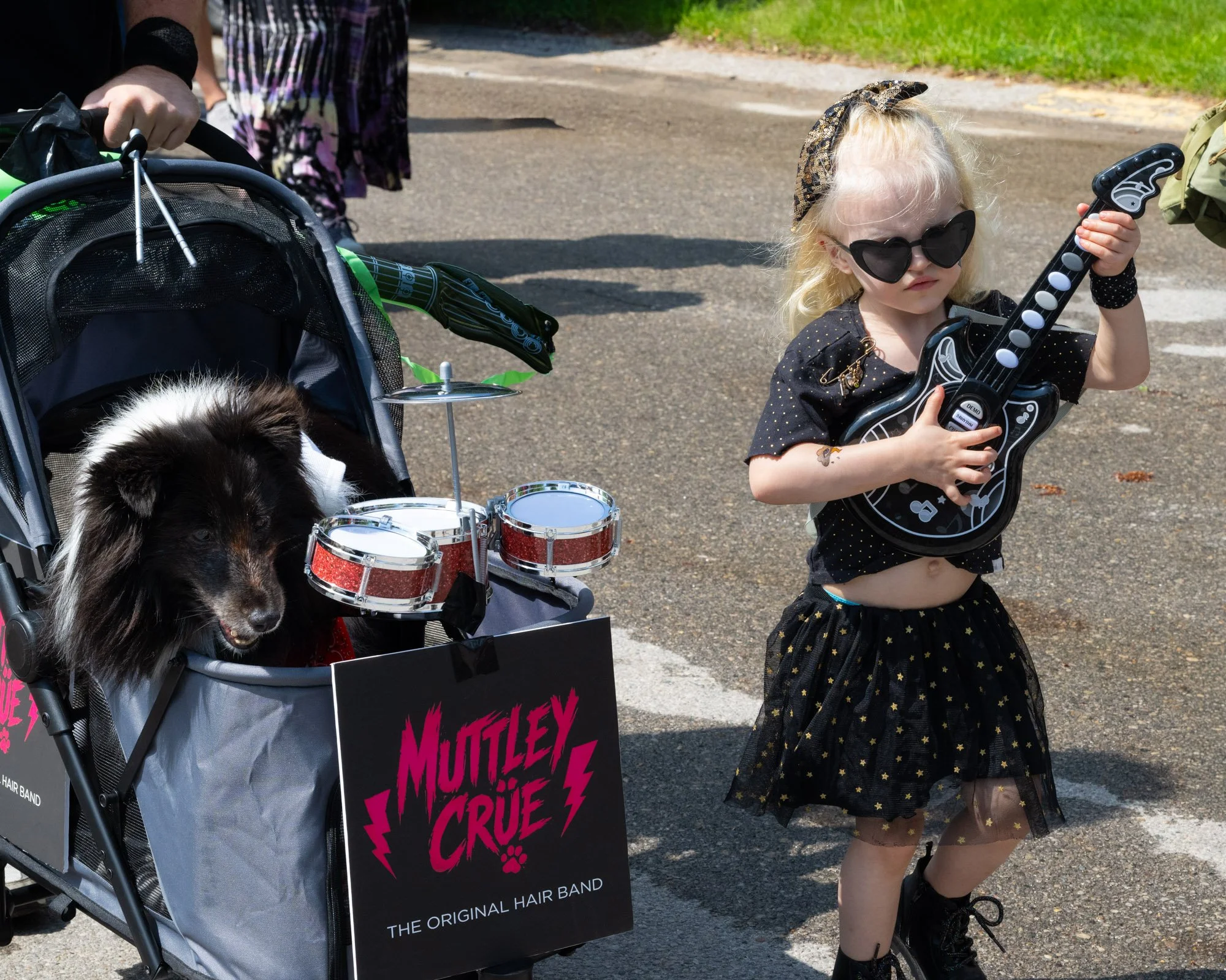 A young girl with blonde curly hair, wearing sunglasses, a headband, and a black and gold polka-dotted dress, holding a toy guitar, standing outdoors near a stroller with a puppy and drums, with a grassy area in the background.
