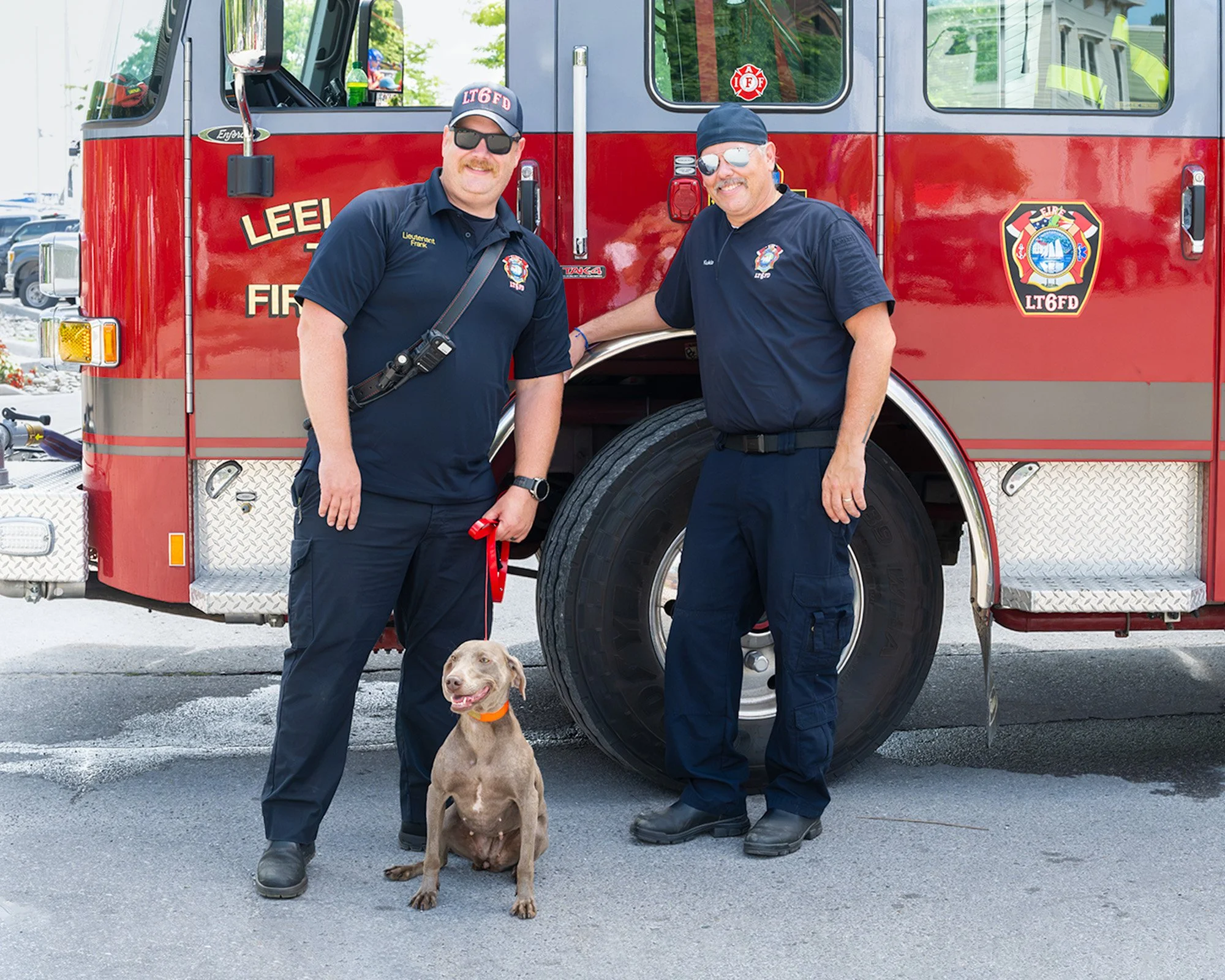 Two firefighters in uniform, one holding a leash with a smiling dog sitting in front of a red fire truck. The firefighters are smiling and wearing sunglasses.