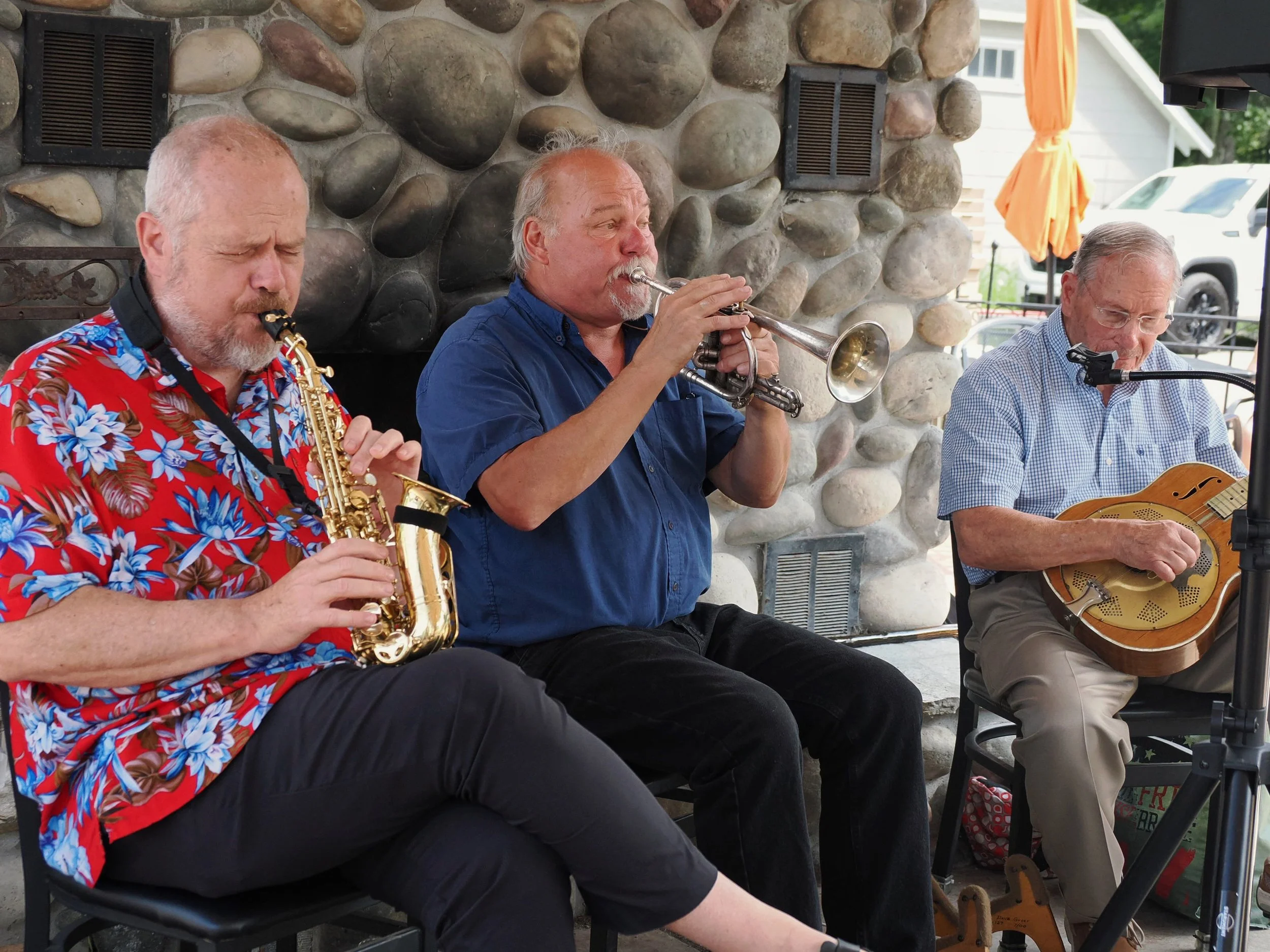 Three older men are sitting outdoors in front of a stone wall, playing musical instruments: one with a saxophone, another with a trumpet, and the third with a guitar.