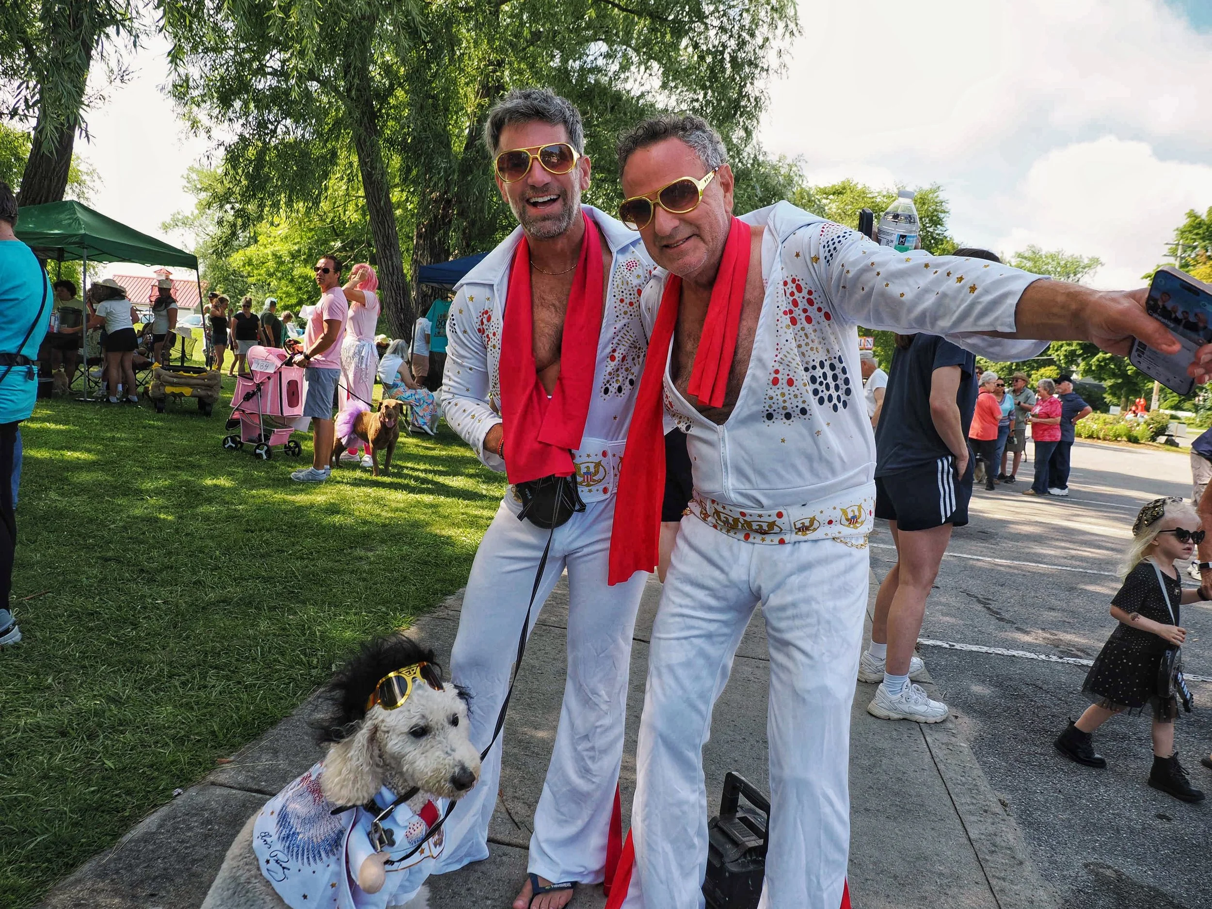 Two men dressed like Elvis Presley posing for a photograph at an outdoor event, with people, tents, and trees in the background.