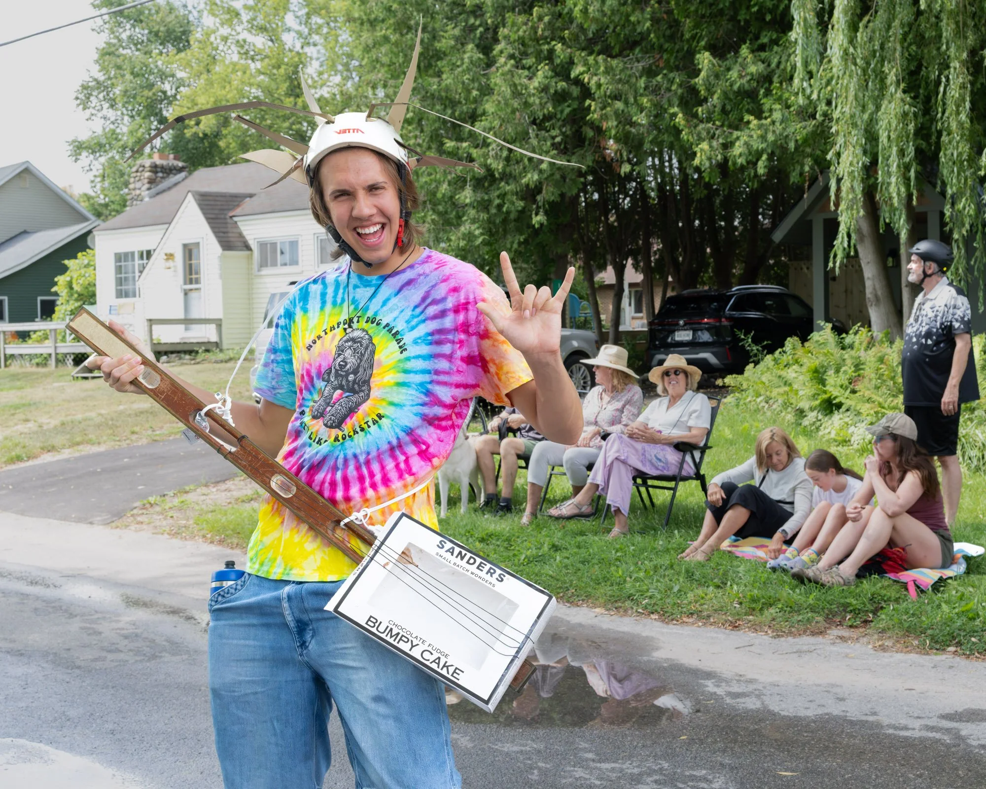 A young man smiling and making a rock on hand sign, wearing a tie-dye T-shirt, a helmet with toy airplane parts, and carrying a sign for a chocolate fudge cake named Bumpy Cake at an outdoor event with seated audience in the background.