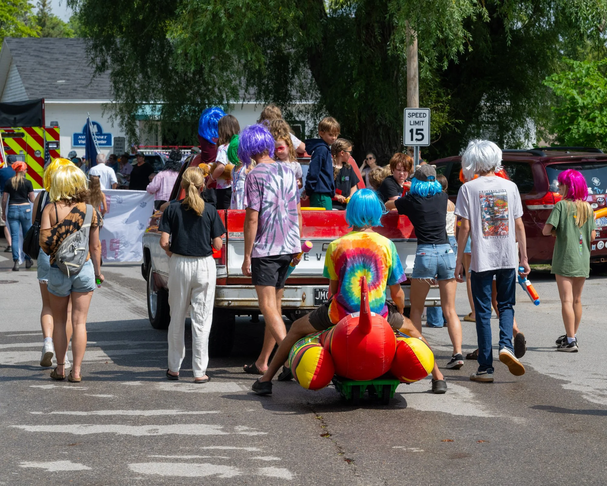 People dressed in colorful wigs and festival attire gather around a small truck with a paddle boat on it, in an outdoor street scene with trees and a speed limit sign of 15 mph.