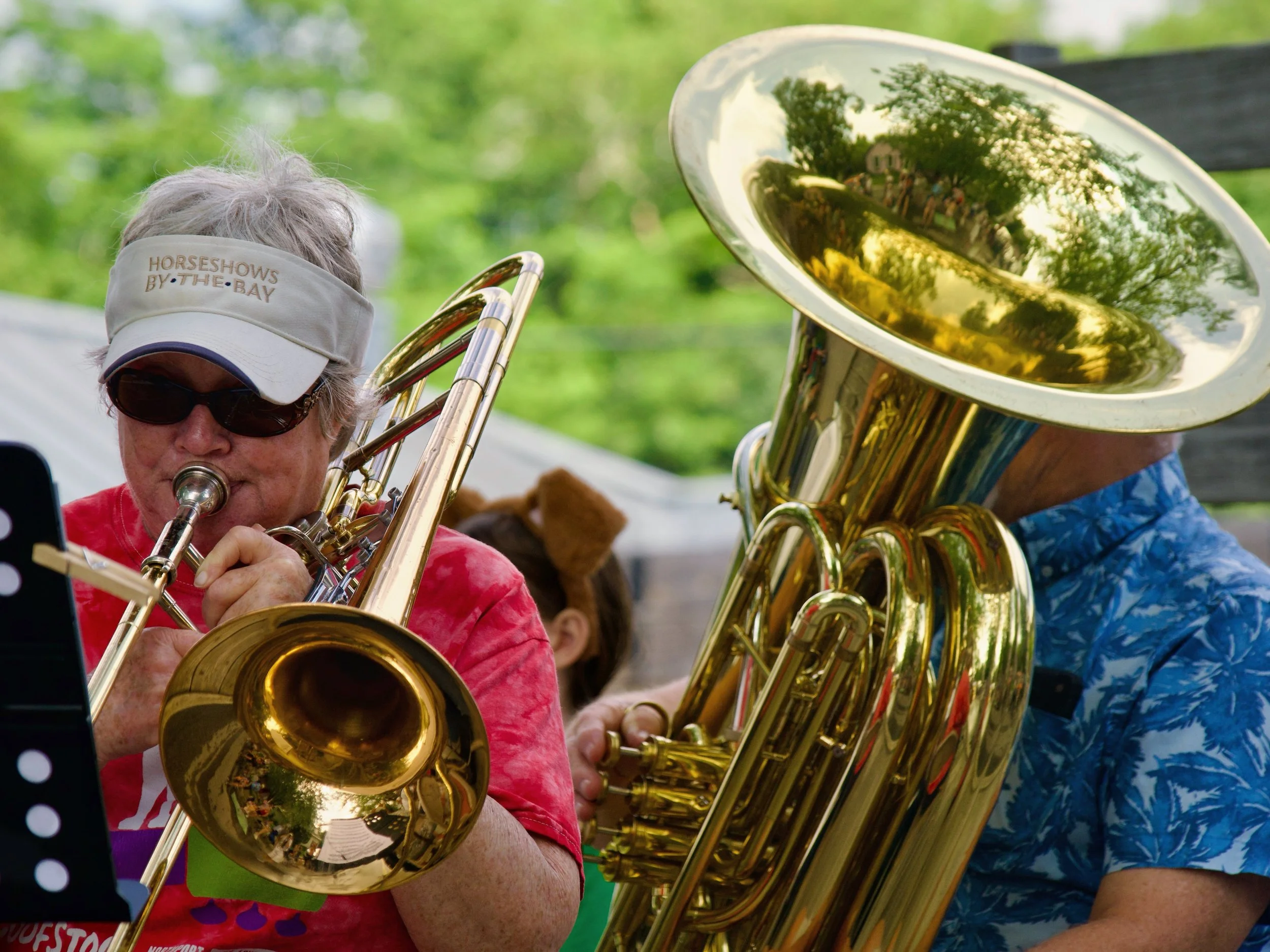 A person wearing sunglasses and a white visor that reads 'HORSESHOES BY THE BAY' playing a brass instrument during an outdoor event, with green trees in the background.