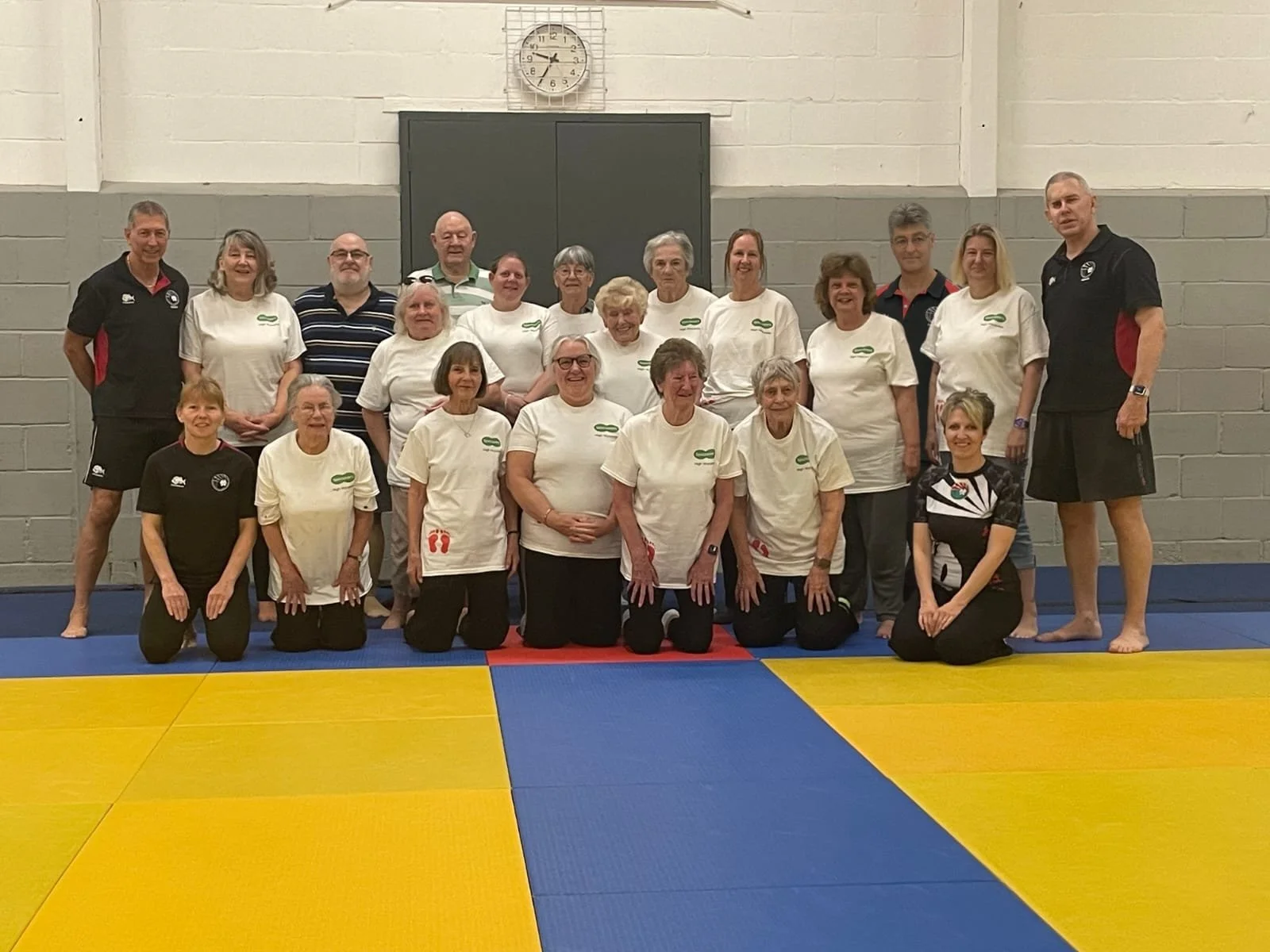 Group of 19 people, mostly women, posing together in a gym with yellow, red, and blue mats. Many are wearing white T-shirts, and some are kneeling in front while others stand behind. Two men in black sports attire stand at each end of the group. A clock showing 8:30 is on the wall behind them.