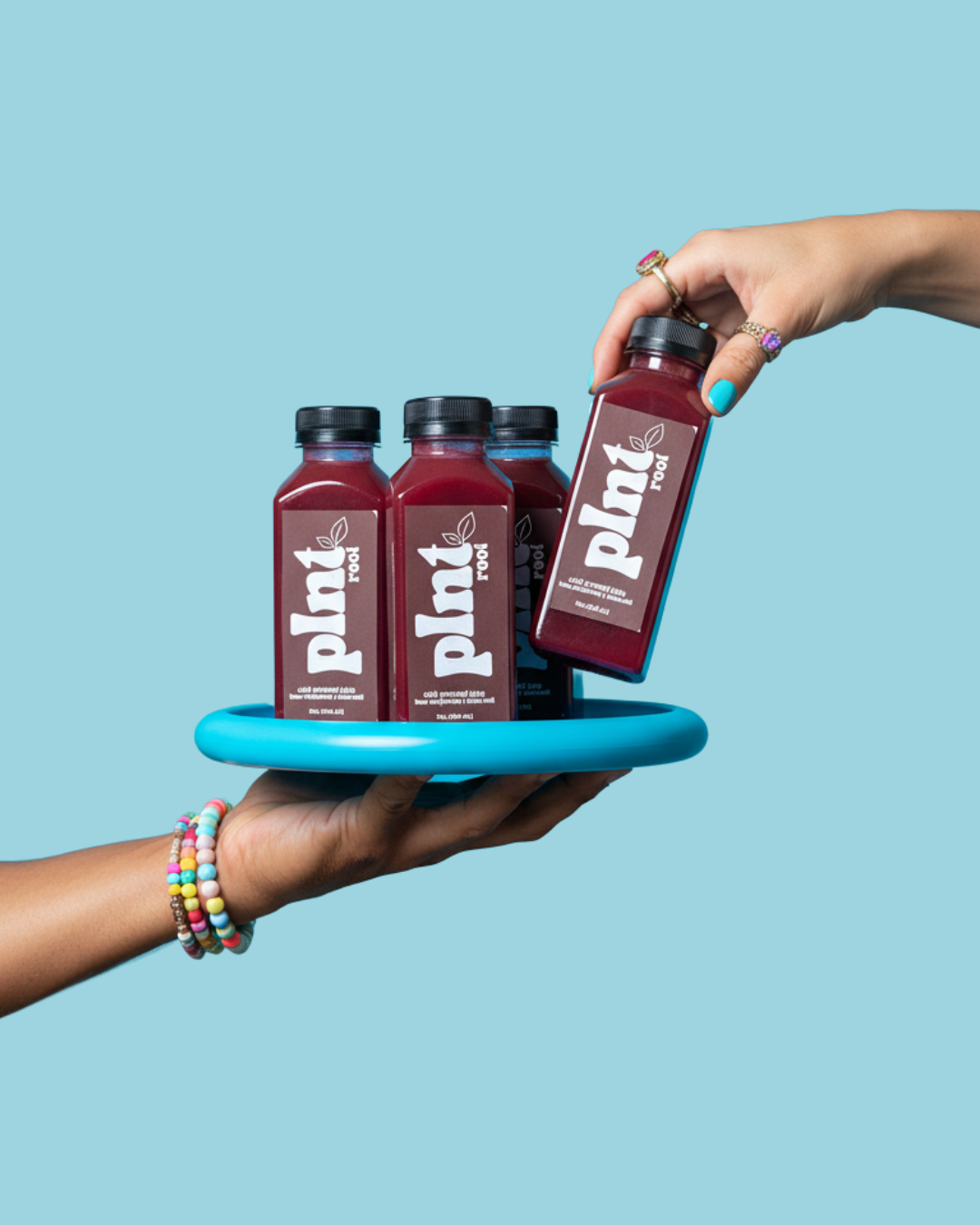 A person with colorful bracelets on their wrist is holding a blue tray with four bottles of plant-based juice labeled "plant root" against a light blue background.