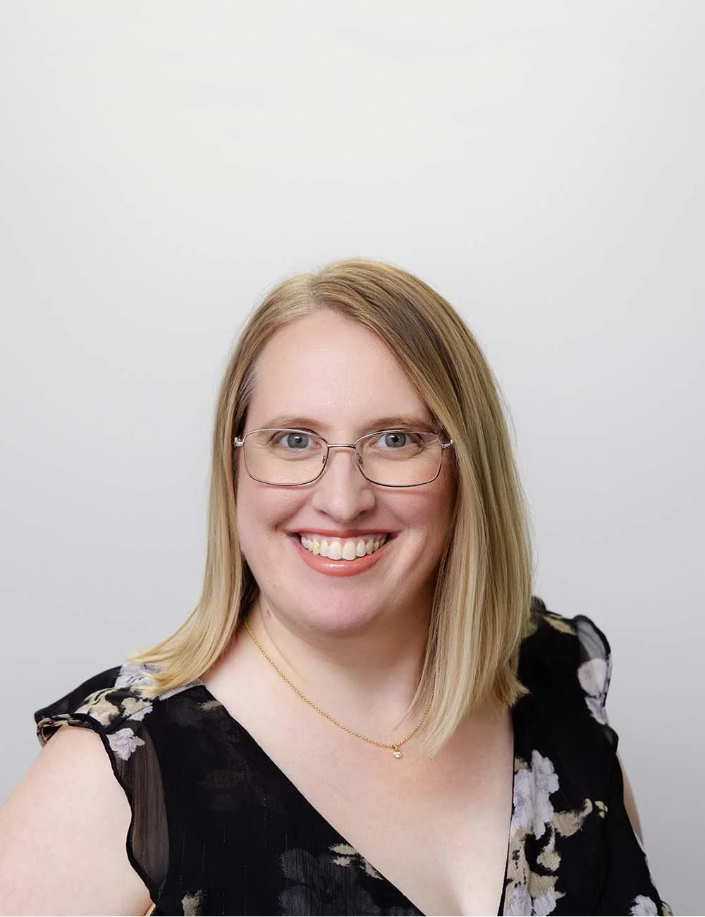A smiling Rebecca Kingdon, Adviser Support at a financial advice firm in Perth. She is a woman with blonde hair, glasses, and a black floral top, standing against a professional white background.