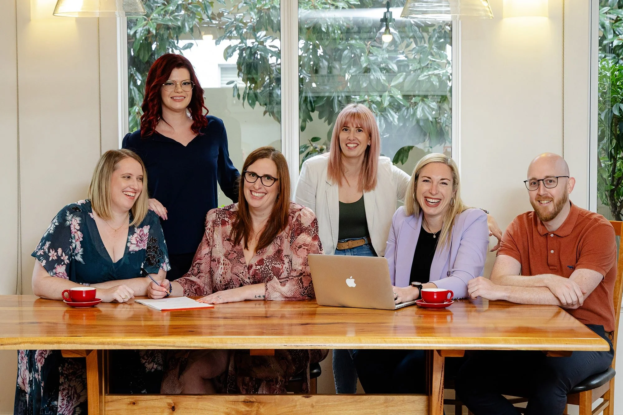 The diverse Firefly Financial team gathered around a wooden table in a meeting room, smiling for the camera. The table has a laptop, notebooks, and red coffee cups.