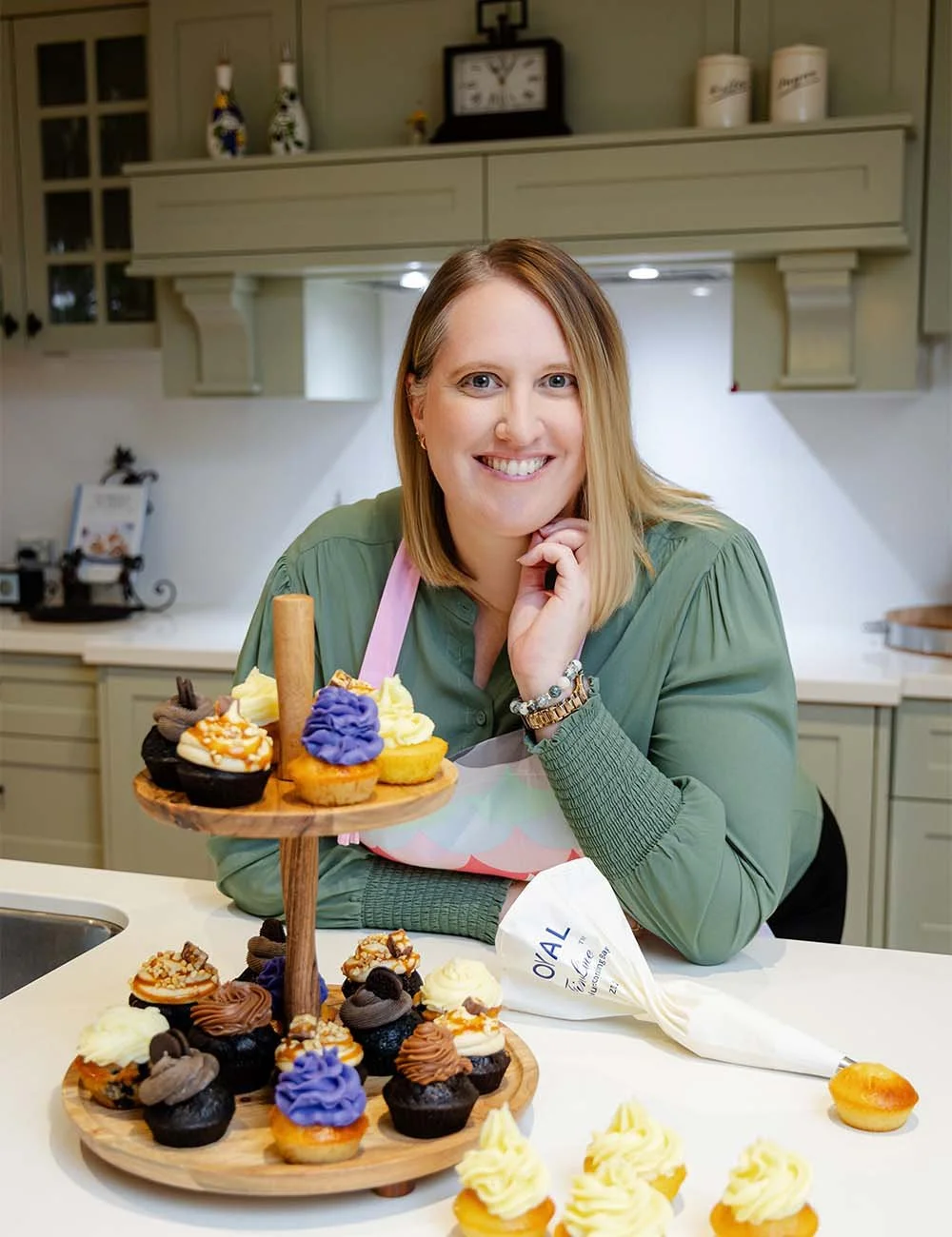 Rebecca Kingdon smiling with shoulder-length blonde hair leaning on a kitchen counter next to two-tiered tray filled with colourful cupcakes and frosting.