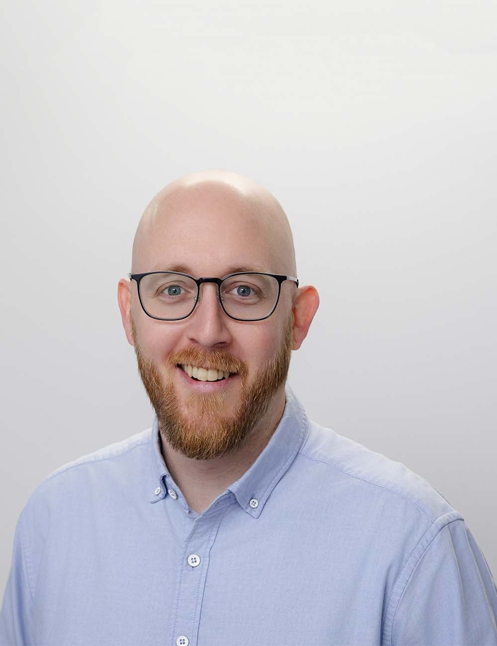 A smiling Scott McGhie, Adviser Support at a financial advice firm in Perth. He wears glasses, a beard, and a light blue dress shirt standing against a white background.