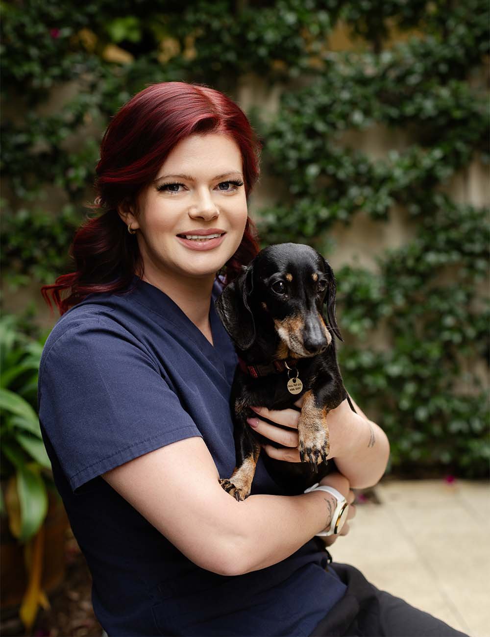 Katie Biggar with red hair wearing navy scrubs holding a black and tan Dachshund dog outside with greenery in the background.