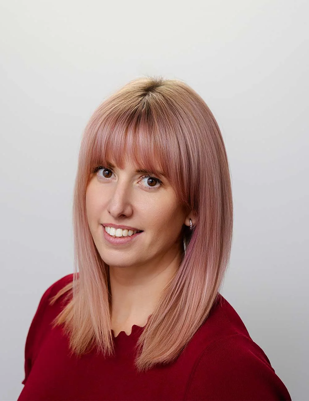 A woman with shoulder-length strawberry blonde hair and bangs smiling at the camera. She is wearing a red top and small hoop earrings. The background is plain white.