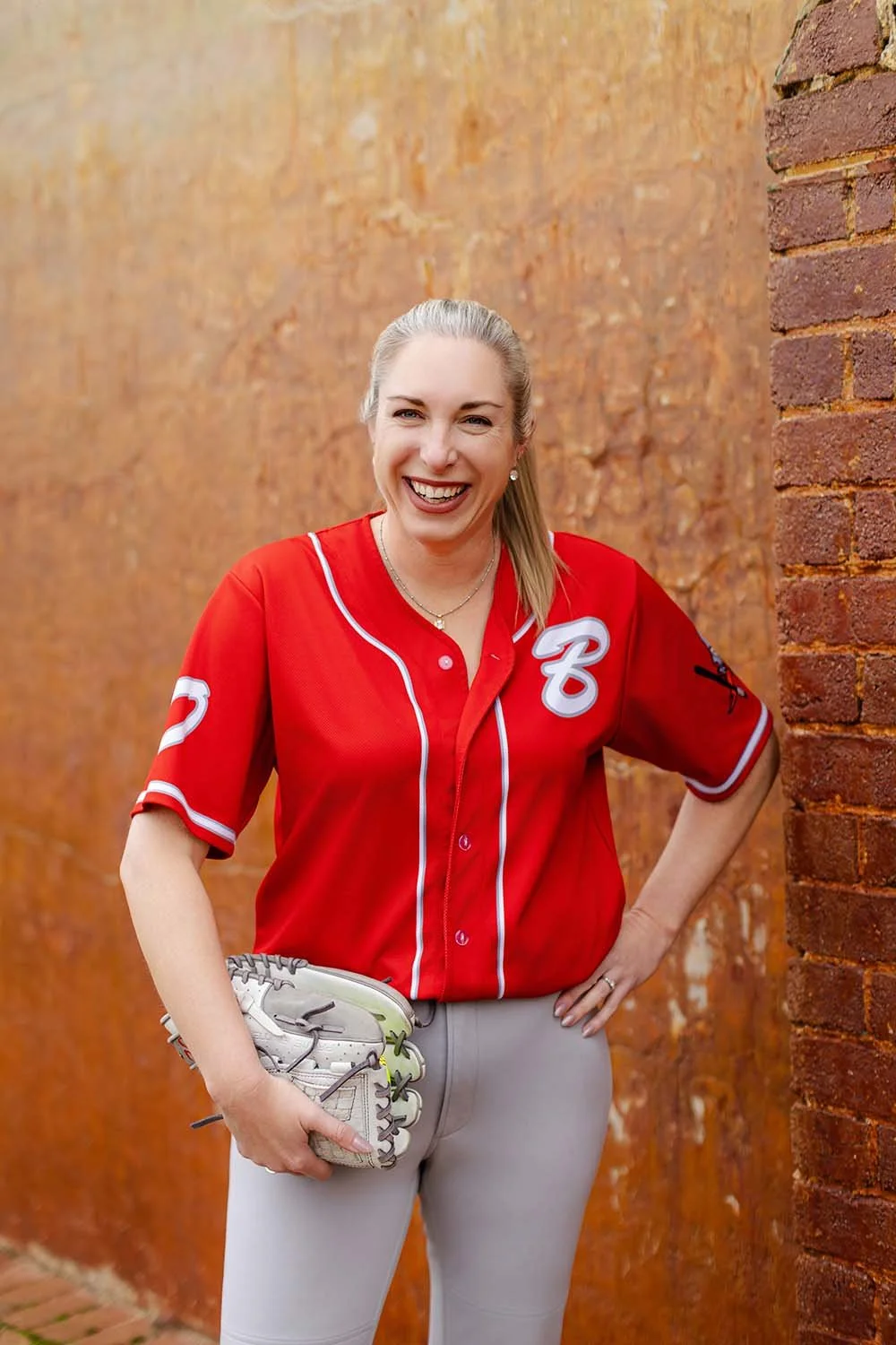Renee Diver smiling and holding a baseball glove, dressed in a red baseball jersey, standing outdoors against a red brick wall.  Financial Adviser by trade, but also plays Australian Masters Games in softball.