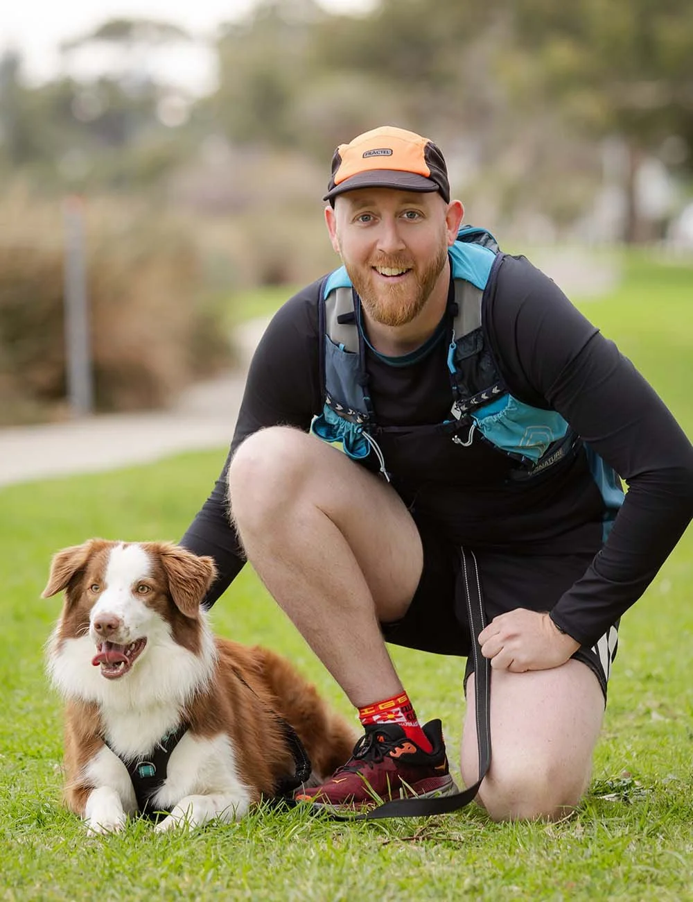 Scott McGhie with a beard, wearing a black athletic shirt, shorts, a backpack, and a colorful cap, kneeling on green grass next to a brown and white Australian Shepherd dog with a harness, in a park with trees and a path in the background.