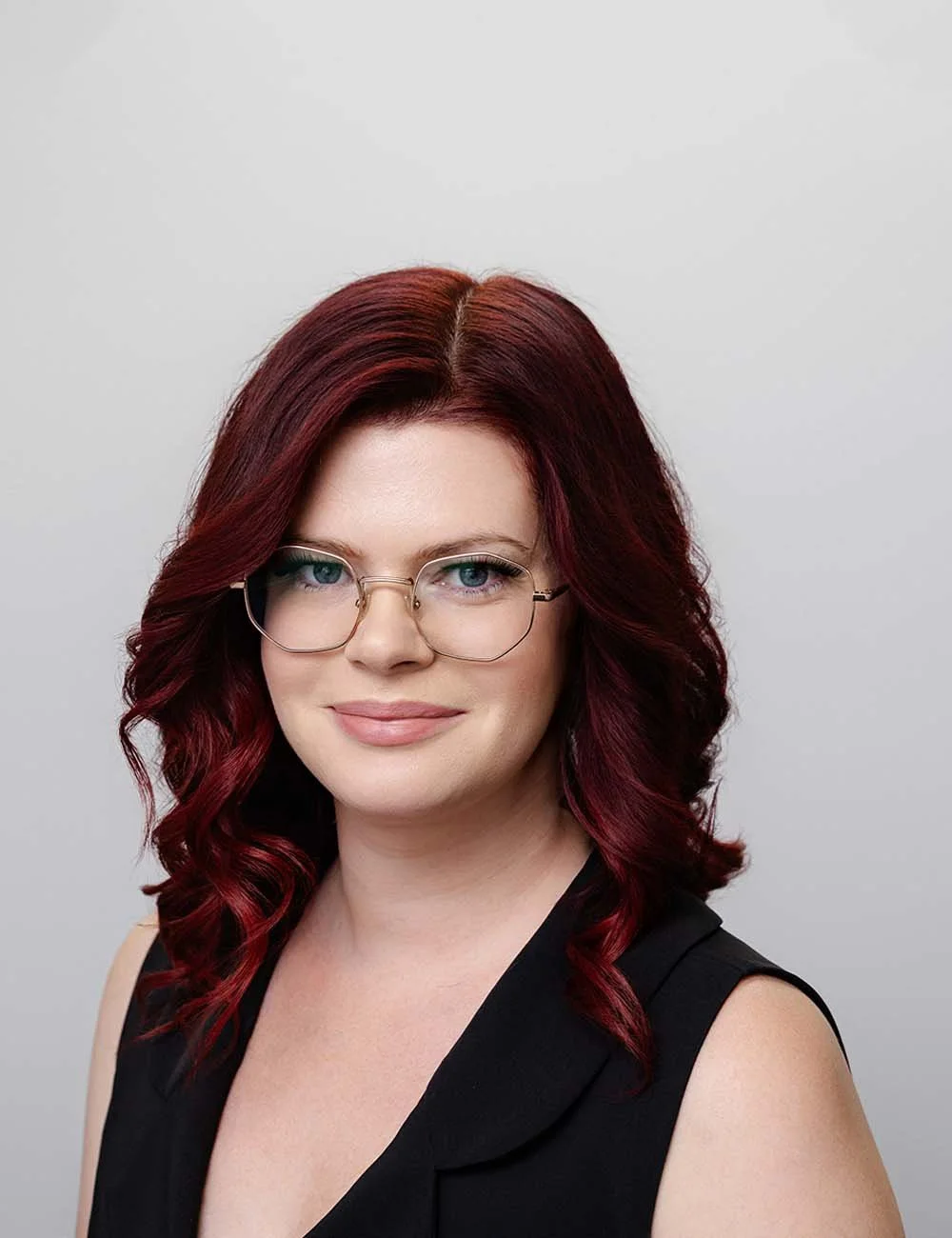 A smiling Katie Biggar, Adviser Support at a financial advice firm in Perth. She has red, wavy hair and glasses smiling at the camera against a professional gray background.