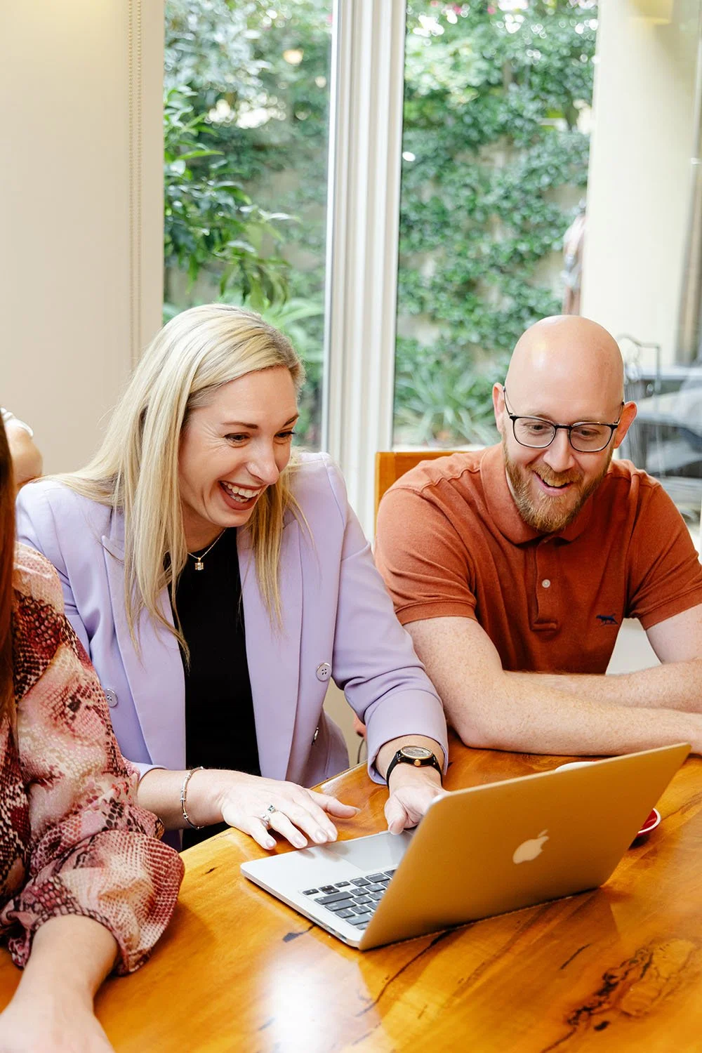 Some of the Firefly Financial team sitting at a wooden table in Cardinya, looking at a laptop and laughing, with a large window and greenery outside in the background.